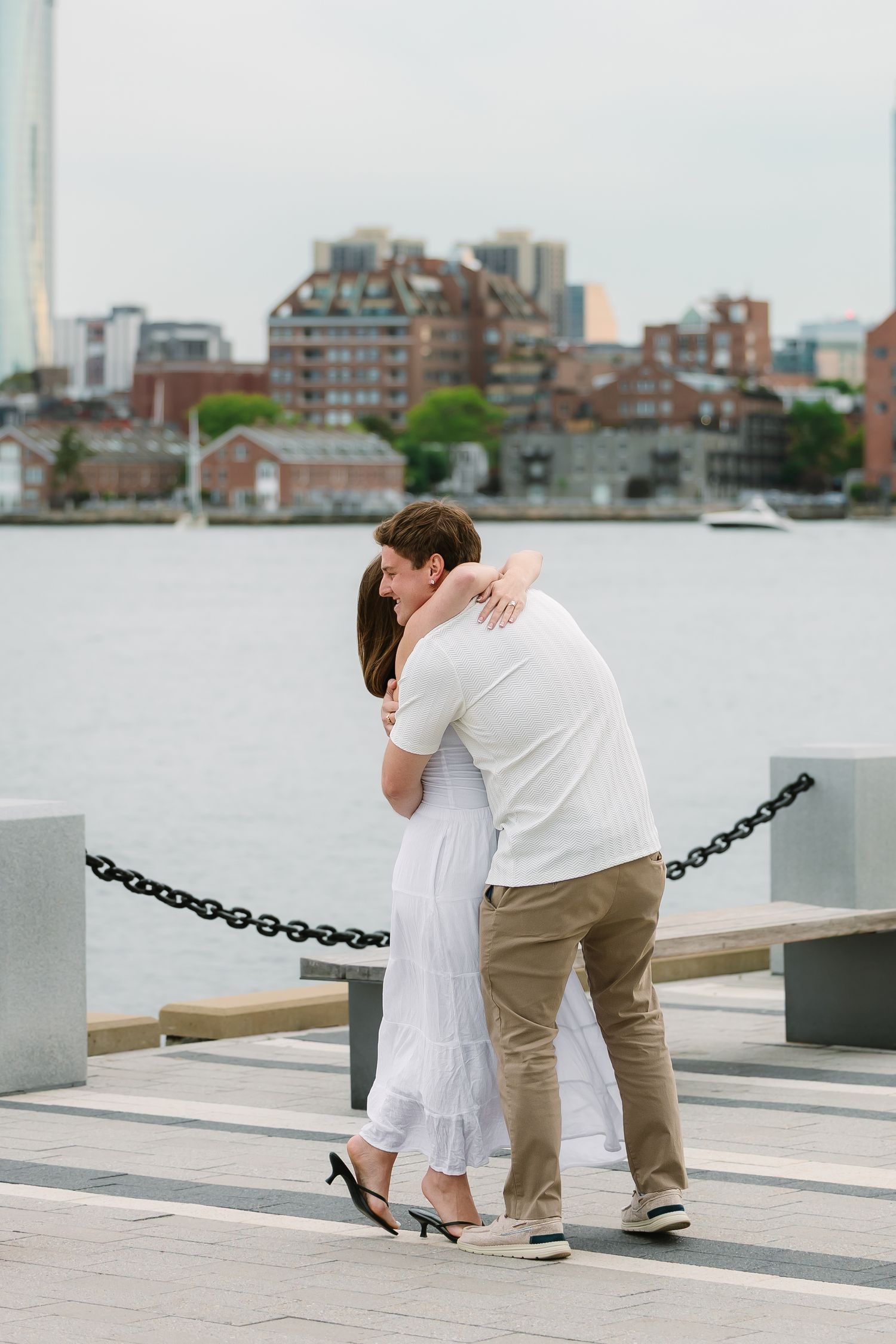 Couple sharing a warm embrace on a waterfront pier with a city skyline behind them.