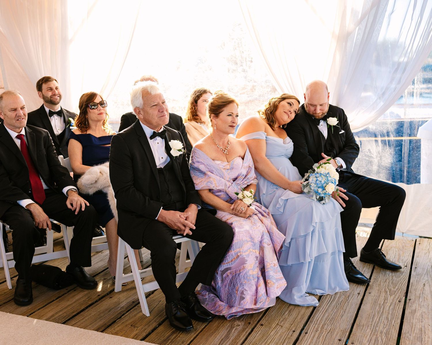 Wedding party members sit together on wooden benches in formal attire at a seaside venue during wedding ceremony at The Regatta Place in RI
