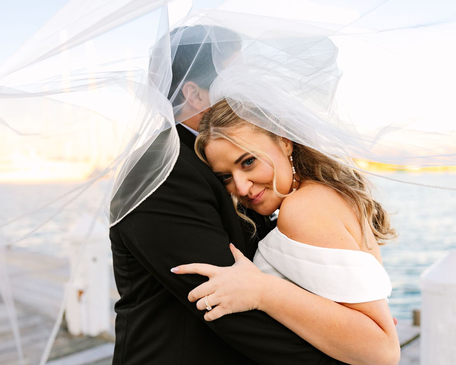 A romantic wedding couple embraces under a flowing veil against a soft sunset sky.