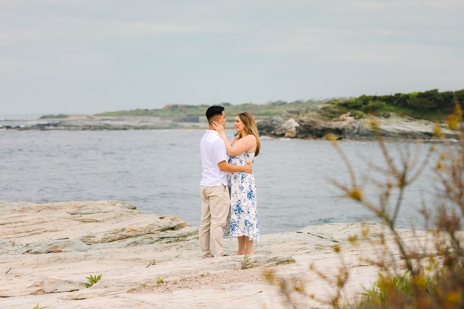 Newly engaged couple embracing on rocky coastal shoreline with ocean views and green foliage on Ocean Drive in Newport, Rhode Island.