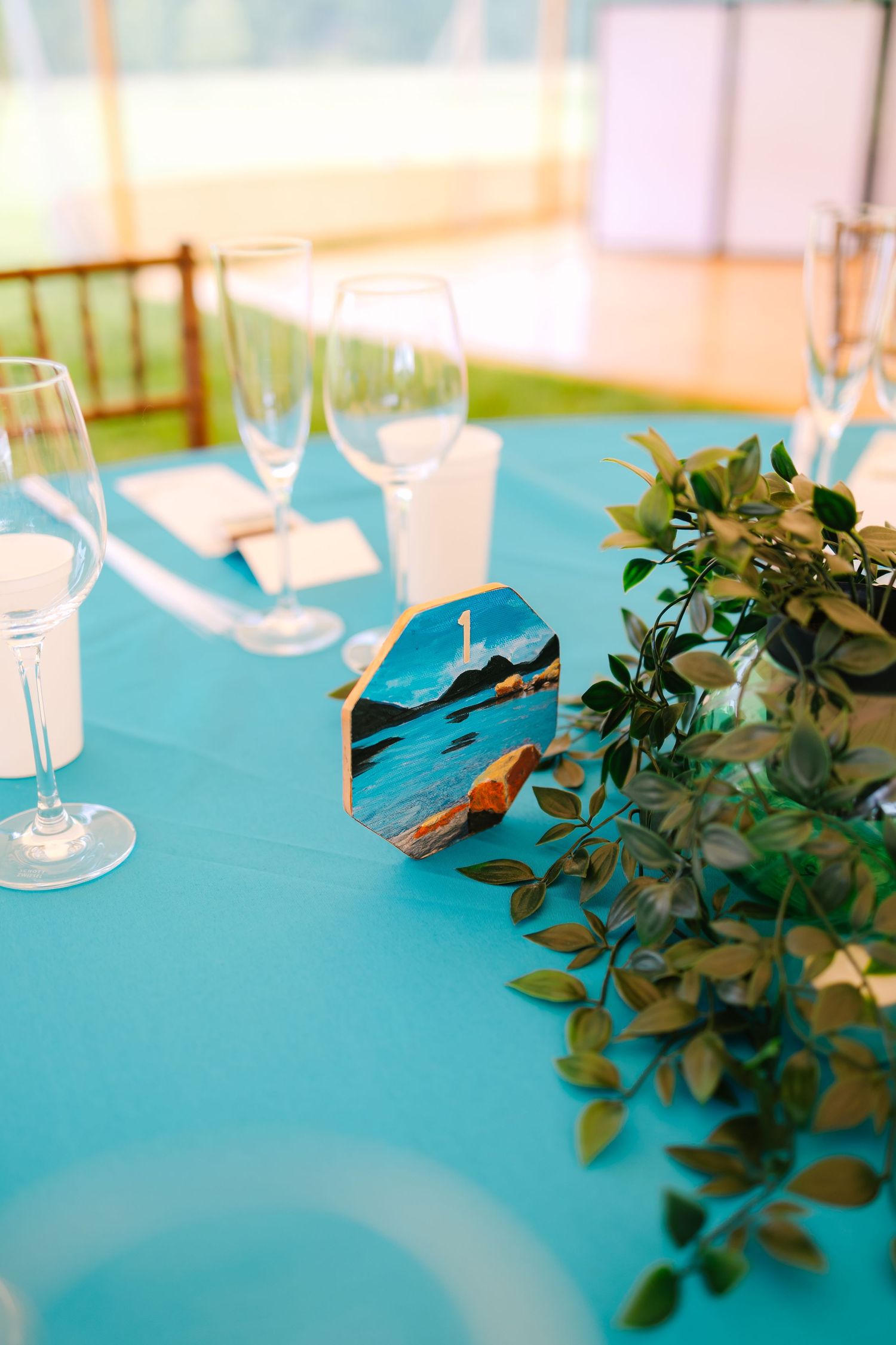 Wine glasses and green foliage decorations arranged on a turquoise tablecloth at an elegant table setting.