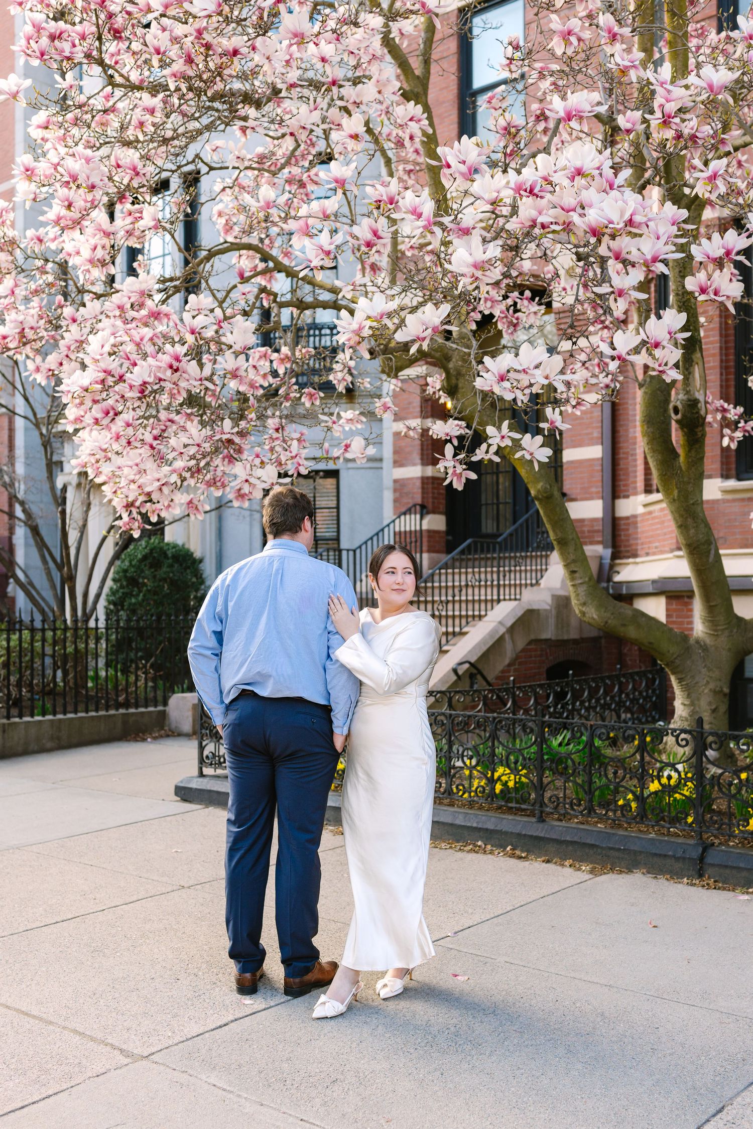 A woman in a white dress embraces her partner beneath a stunning pink magnolia tree in the city.