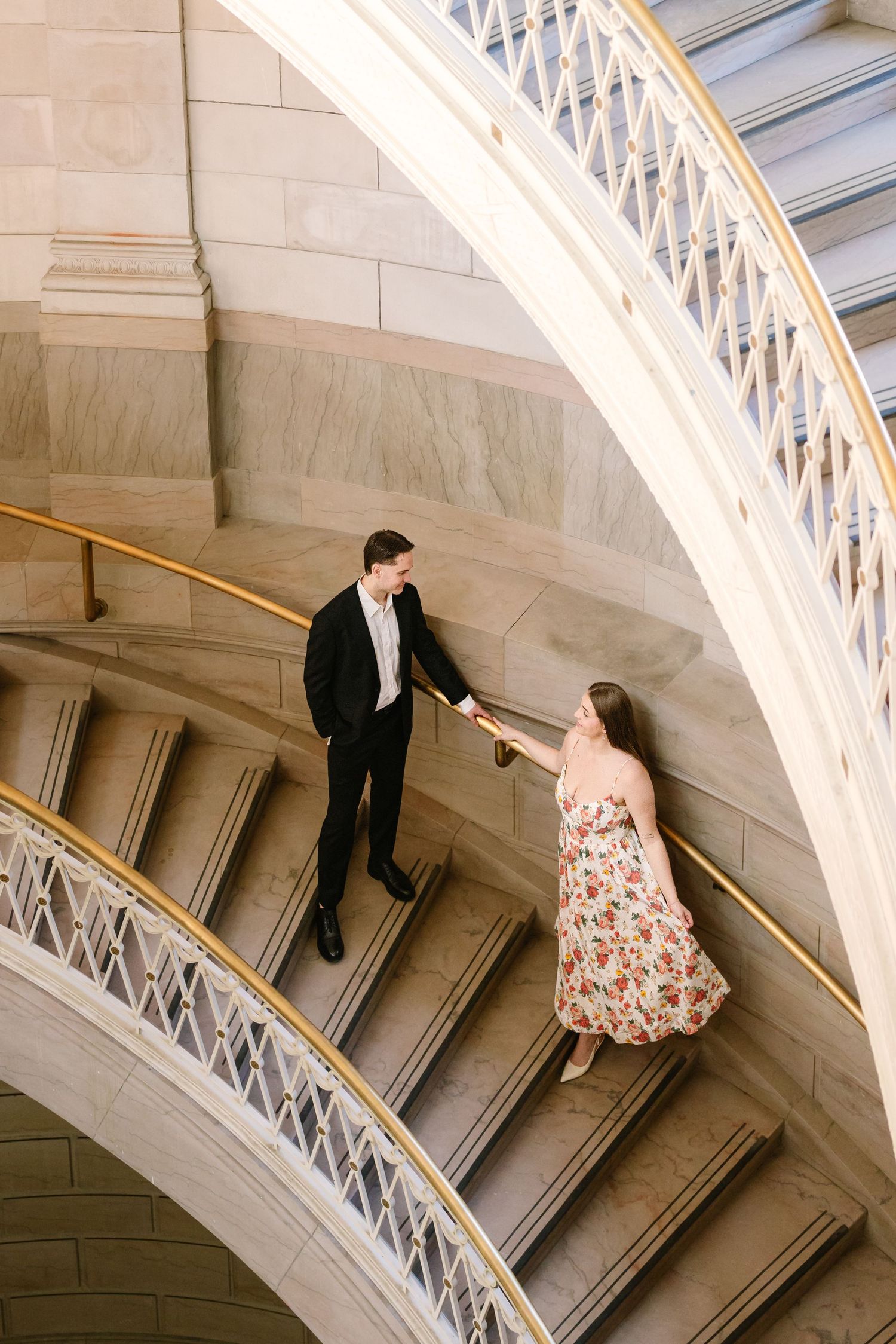 Man in black suit and woman in floral dress holding hands on grand curved staircase with gold railing.