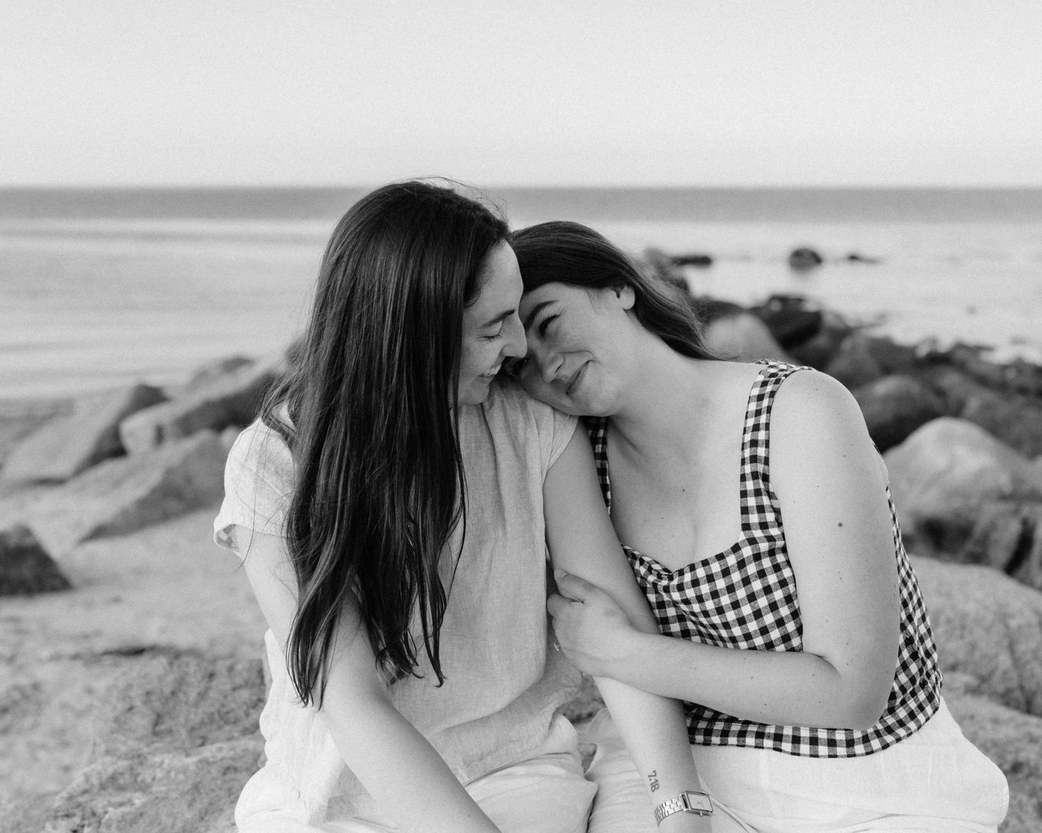 Two women laughing and leaning together on rocky beach in black and white photo.