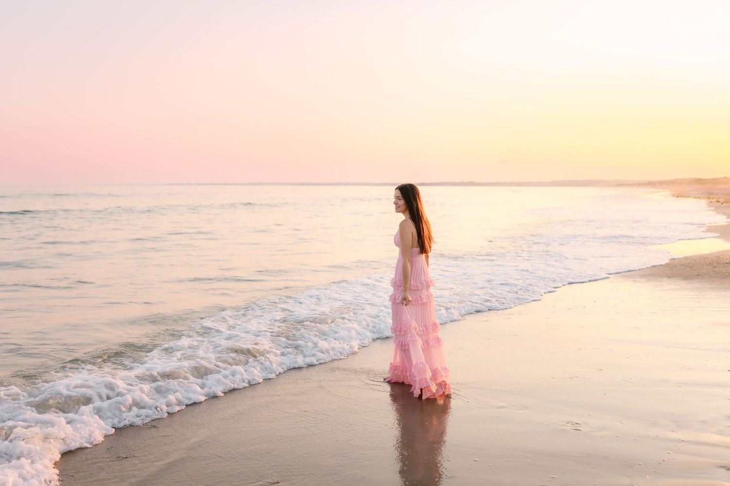 Woman in long pink ruffled dress standing gracefully at the ocean shore during sunset.