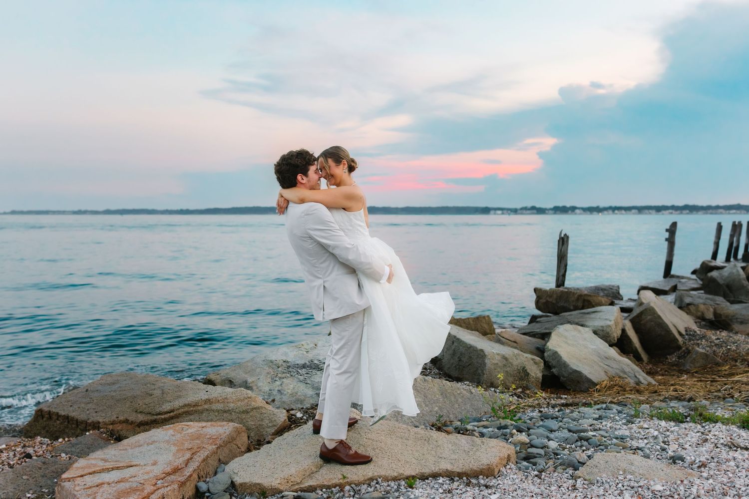Groom lifting bride for a kiss on rocky shoreline with colorful sunset sky behind them.