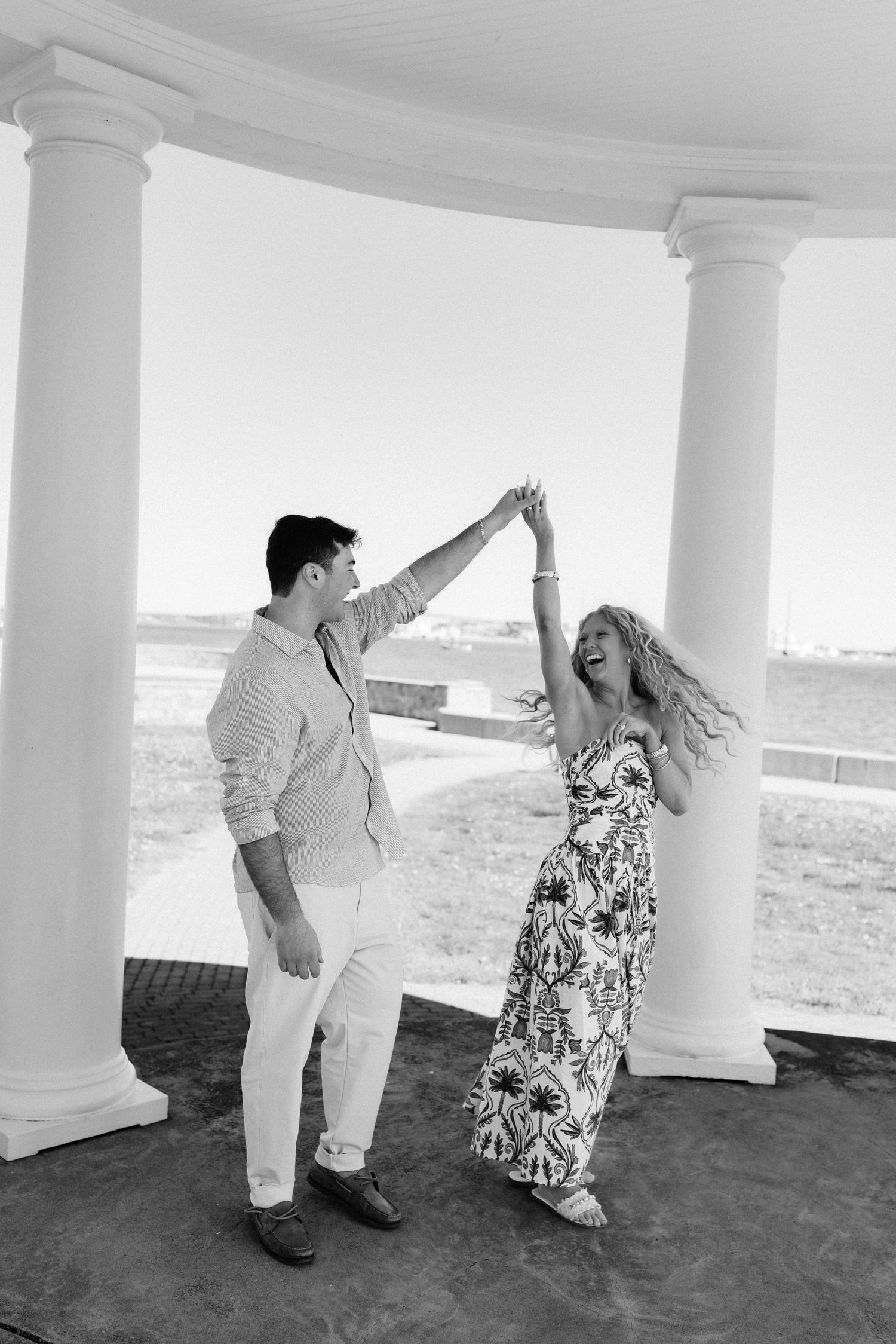 Black and white photo of a joyful couple dancing under a columned gazebo by the waterfront.