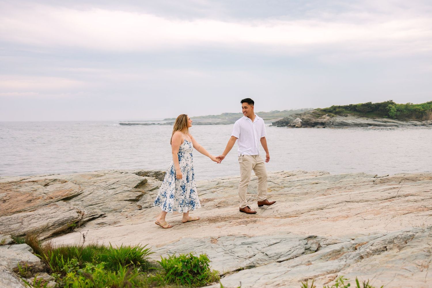 A couple holding hands walking along the coastal rocky cliffs of Newport Rhode Island on Ocean Drive for engagement photos after proposal.