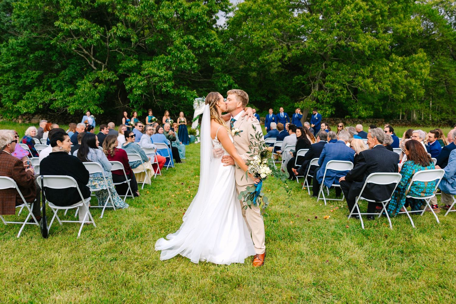 A bride and groom share a kiss at their wedding ceremony recessional with wedding guests in the background for backyard wedding in CT