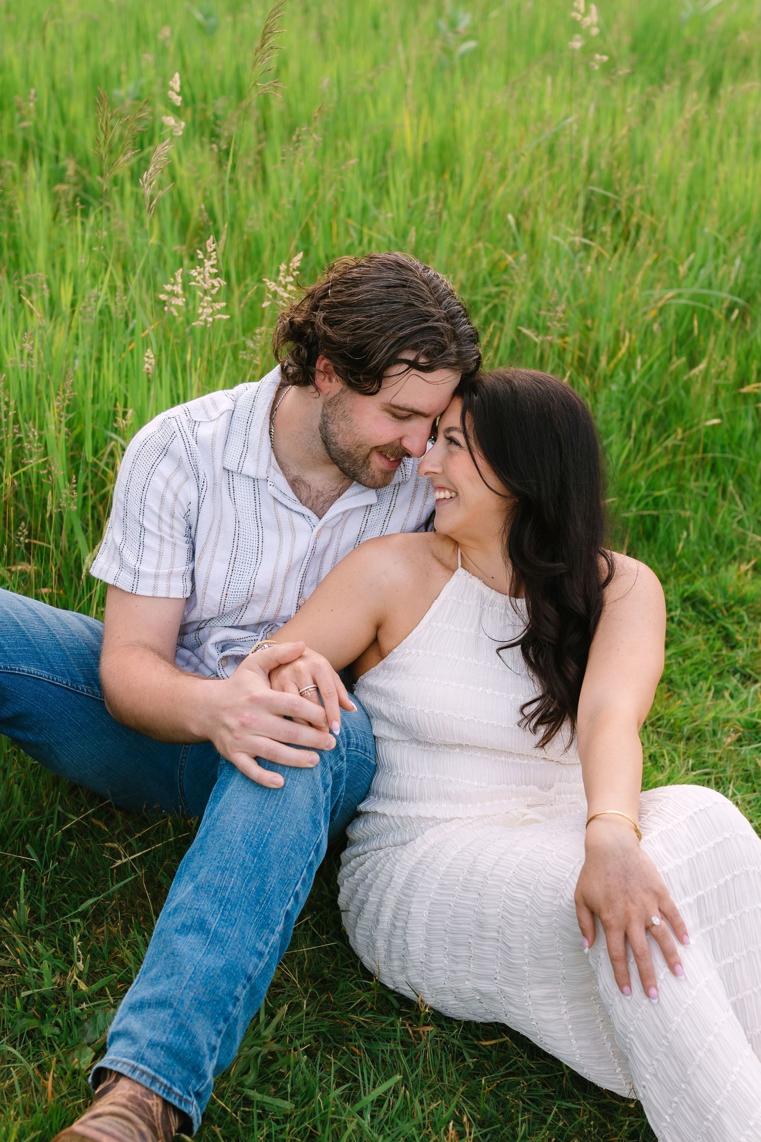 Couple sitting close together in tall green grass, smiling and touching foreheads.