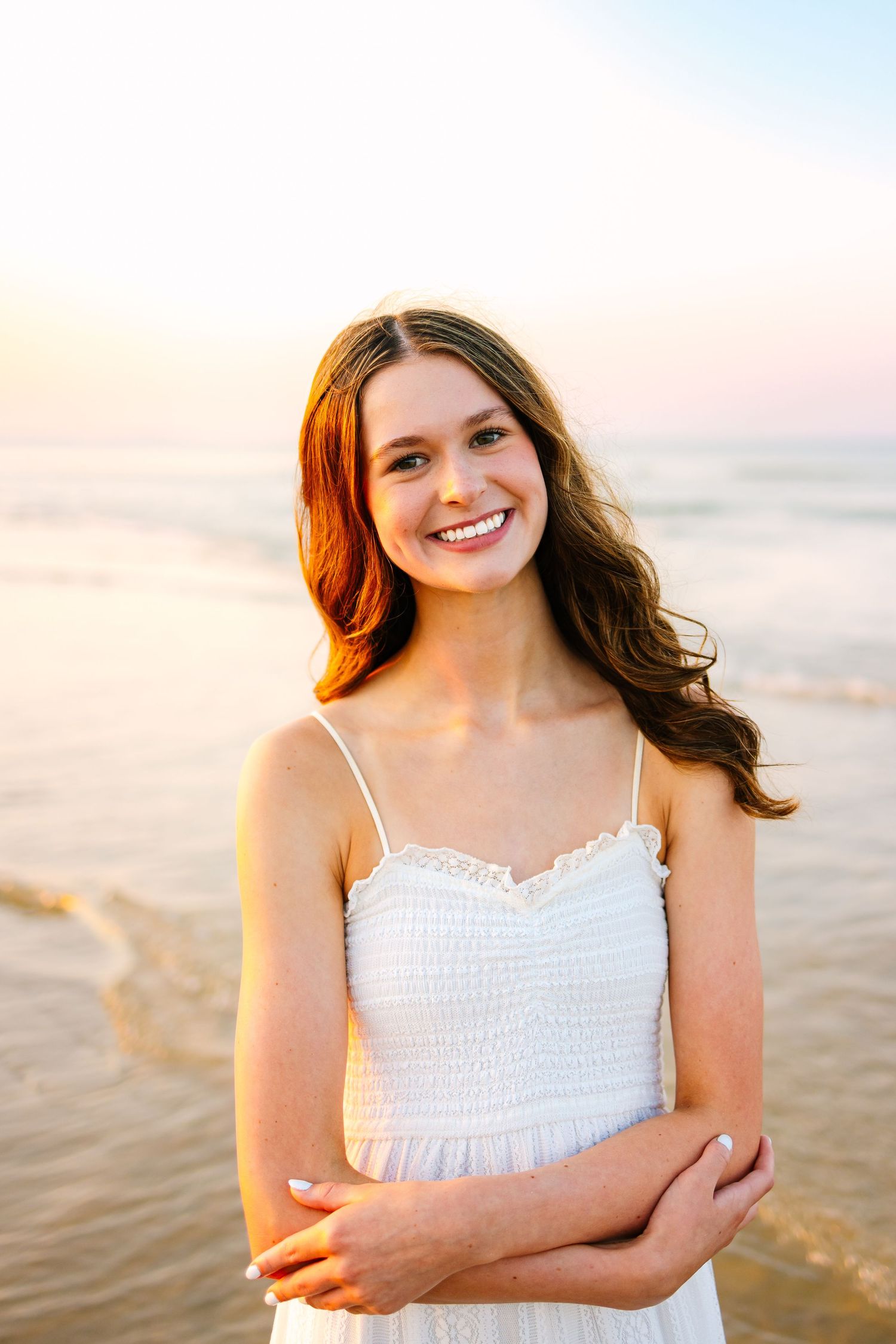 Smiling young woman in white spaghetti strap dress standing on beach at sunset with arms crossed.