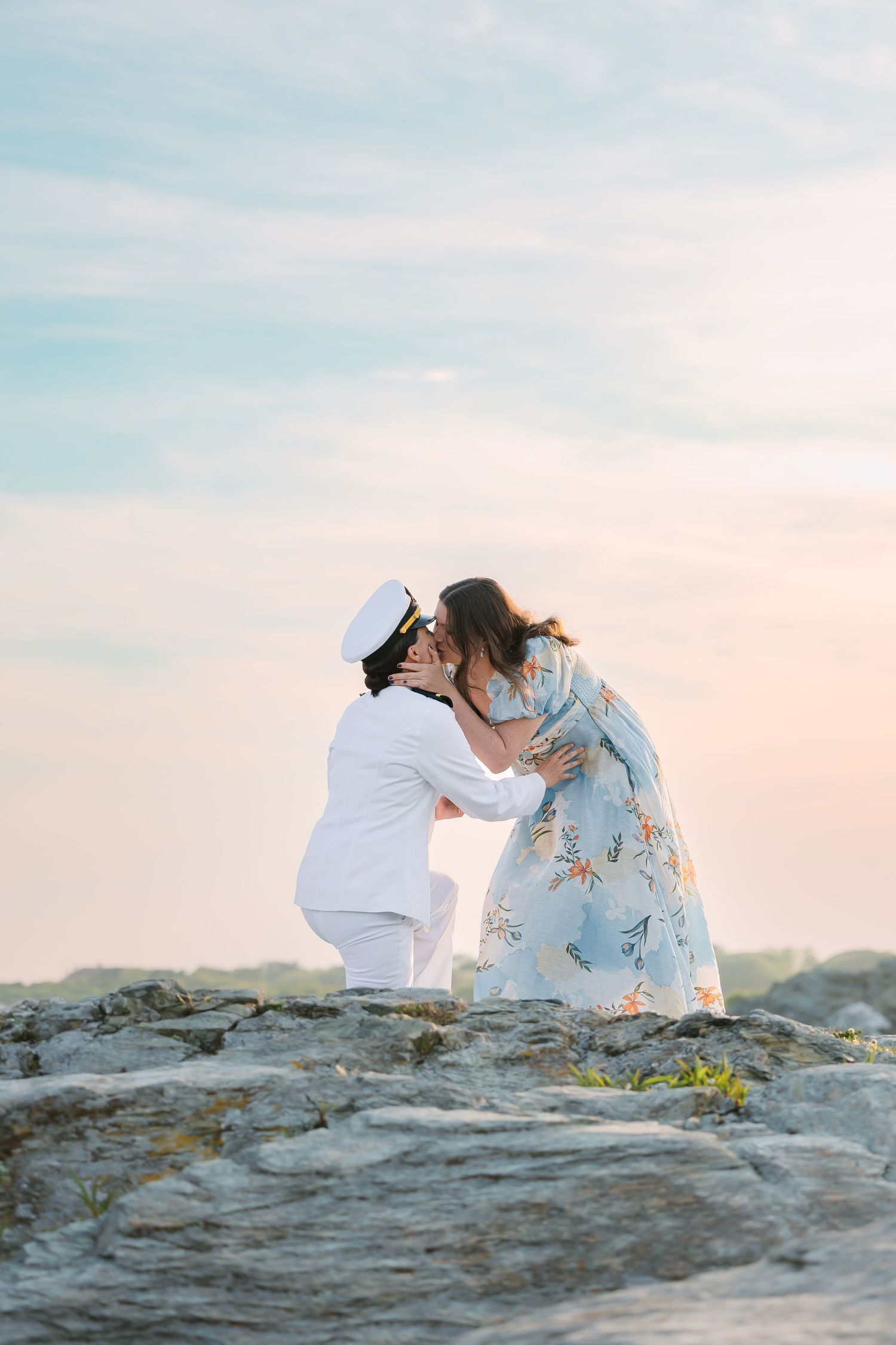 A sailor in white uniform kneels to kiss his partner on rocky coastal terrain at sunset.