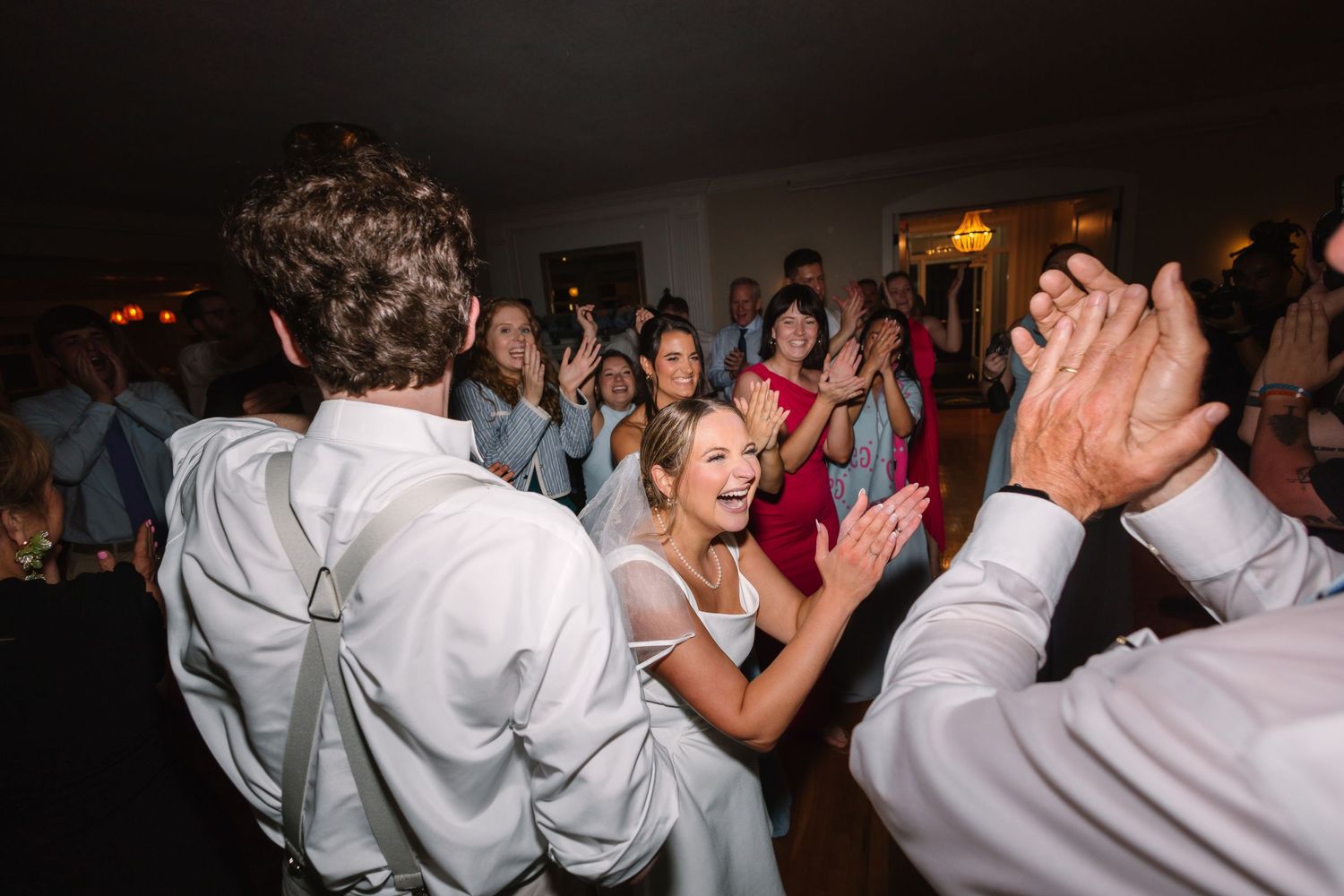 Smiling bride in white gown dances joyfully with guests clapping at lively wedding reception.