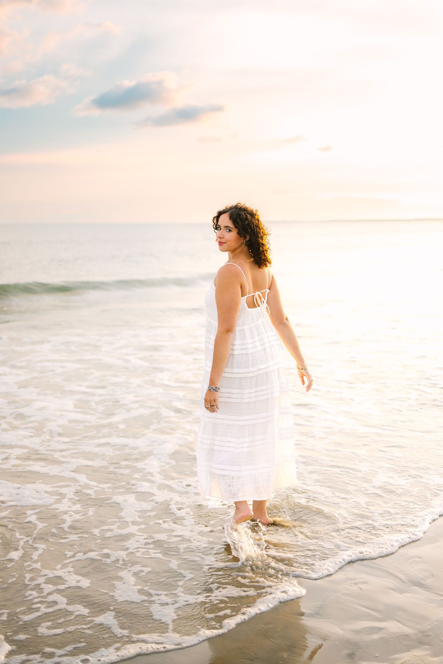 Woman in white maxi dress standing at ocean's edge during a beautiful sunset.