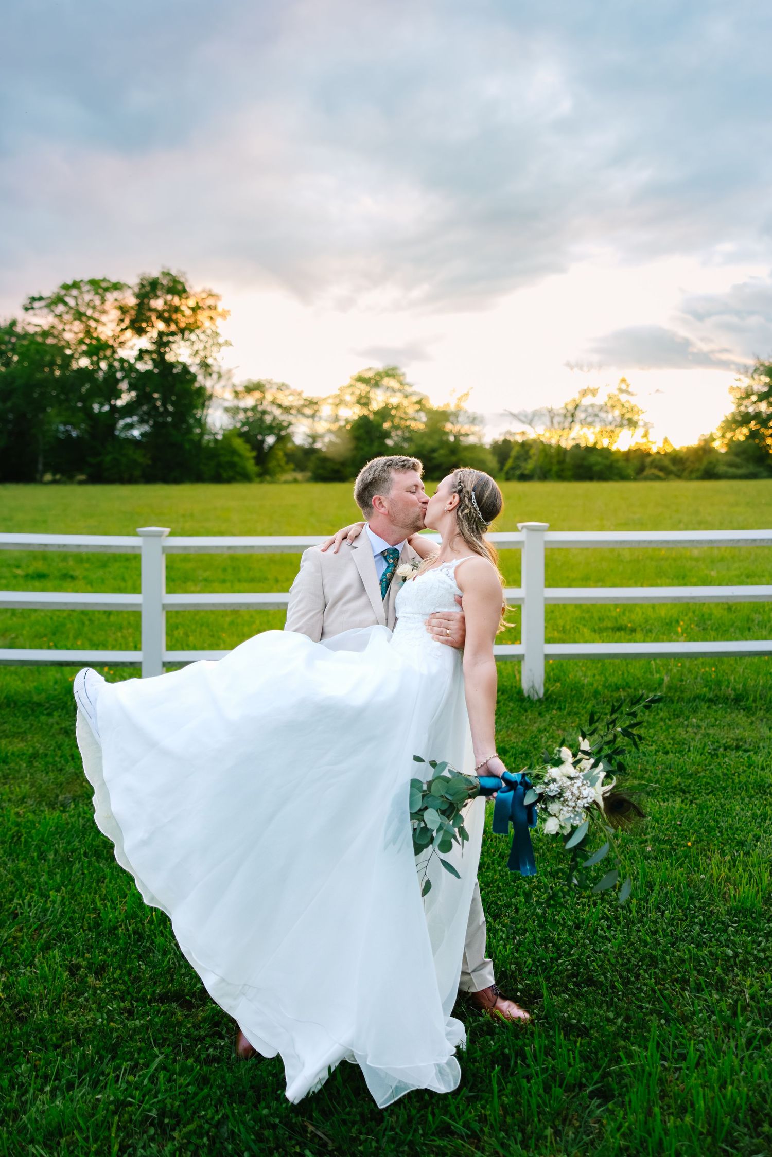 A romantic wedding photo captured at sunset by a white fence with flowing white dress and flowers in a grassy field.