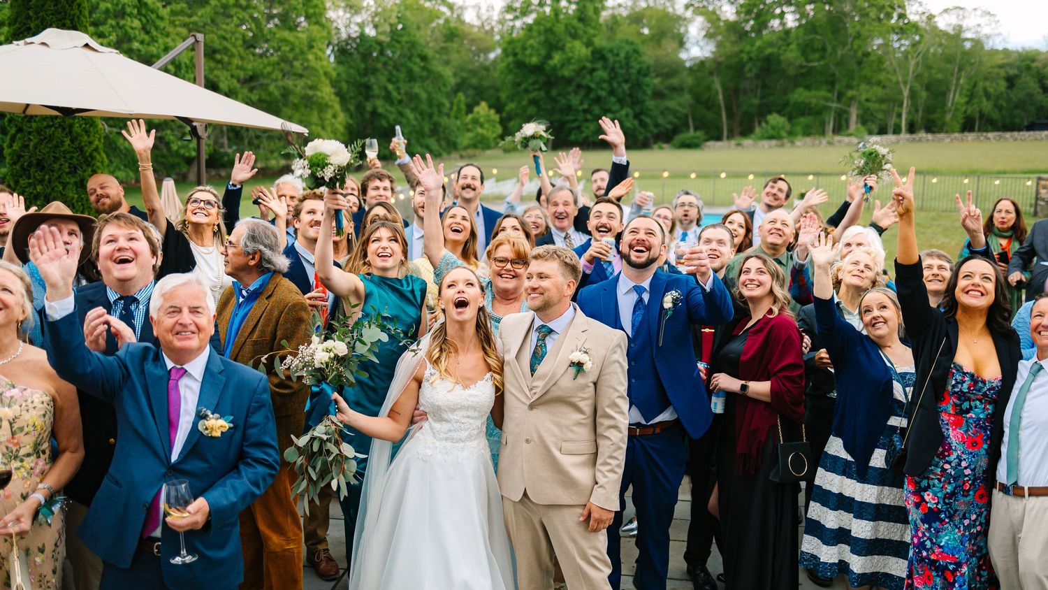 Large wedding party cheers and raises their hands in celebration outdoors on sunny day with green trees in background.