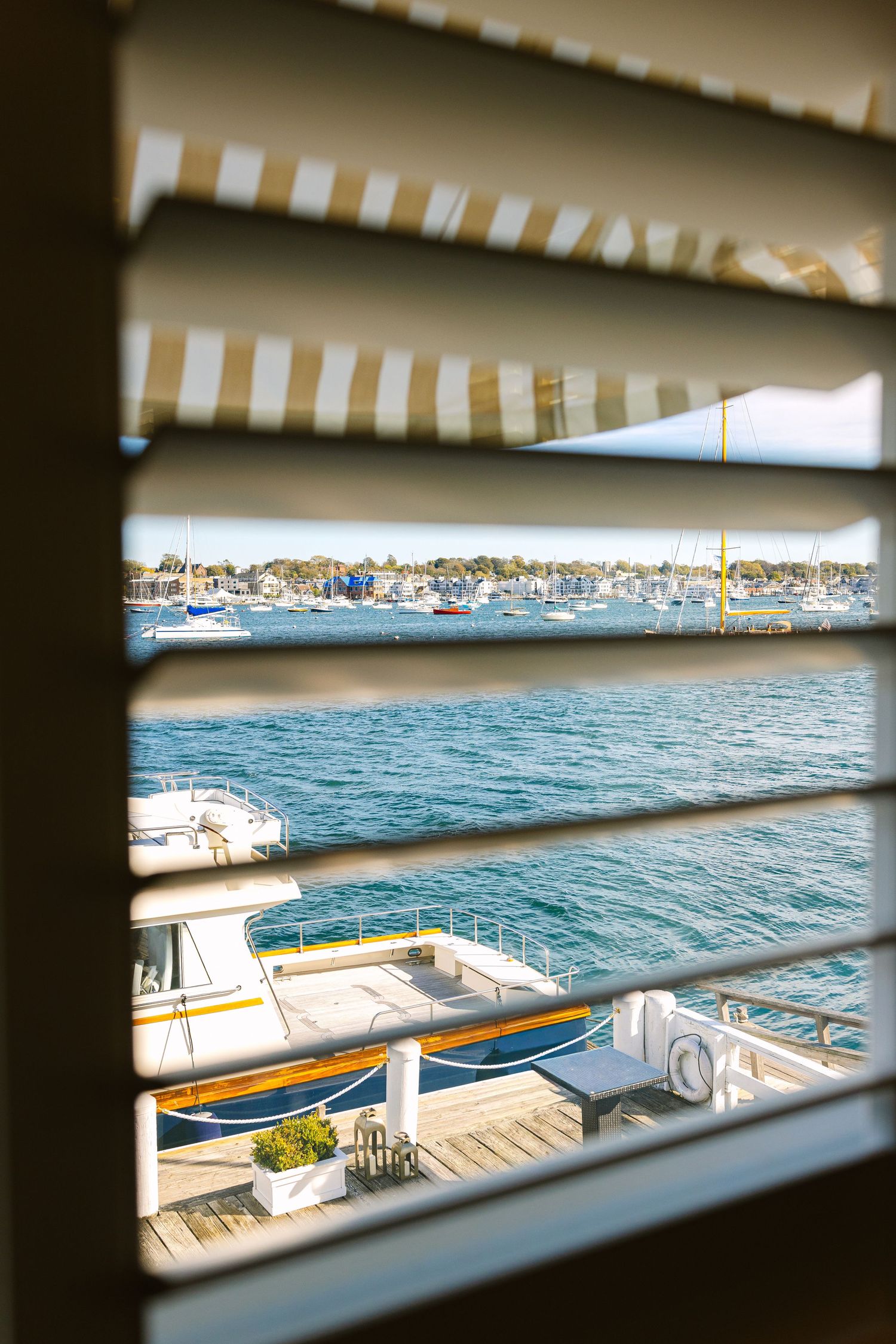 View through window blinds of boats docked in a marina with blue water and coastal scenery in background at The Regatta Place in Newport RI