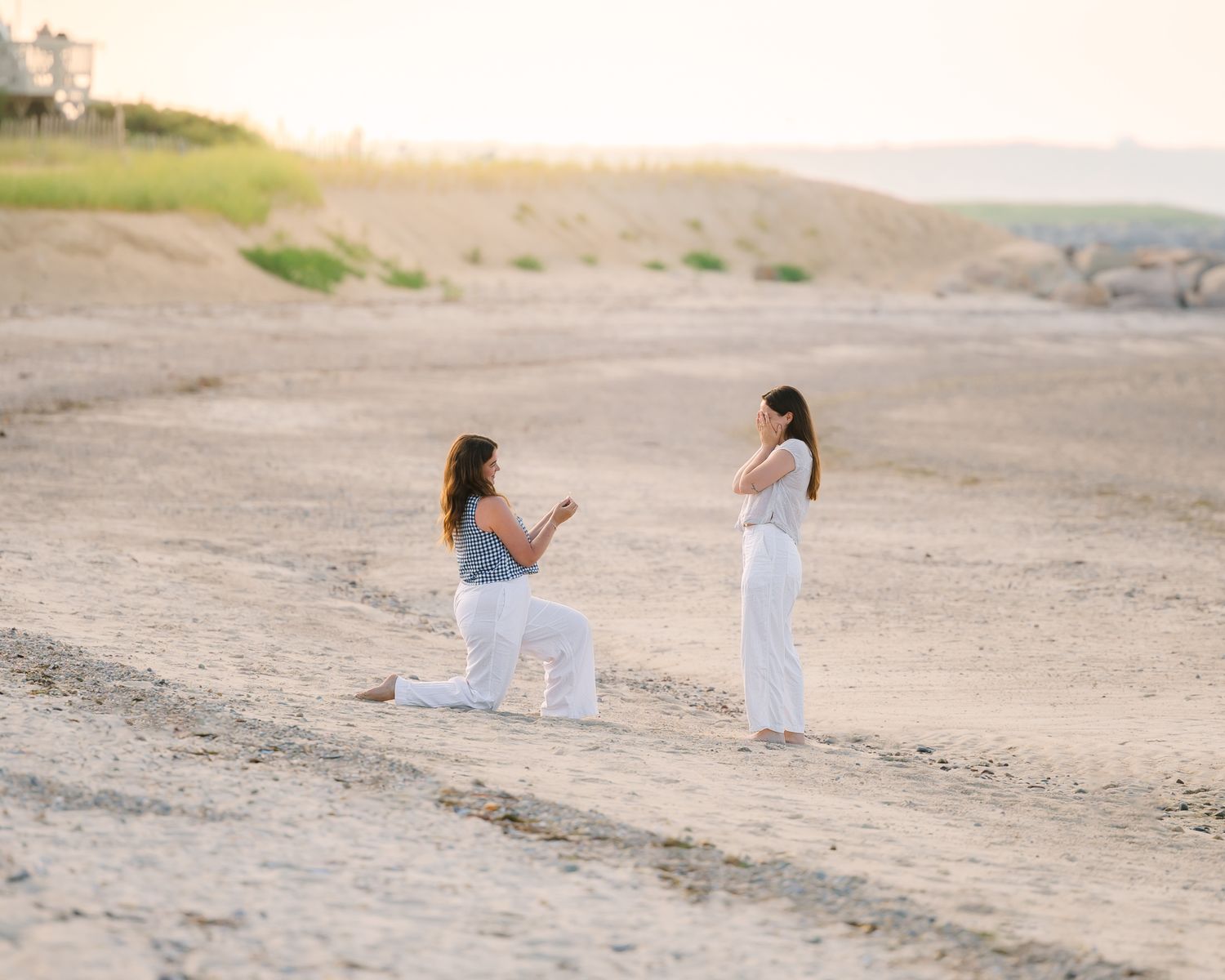Two women in white pants share a beach proposal at sunset, one on bended knee holding a ring.