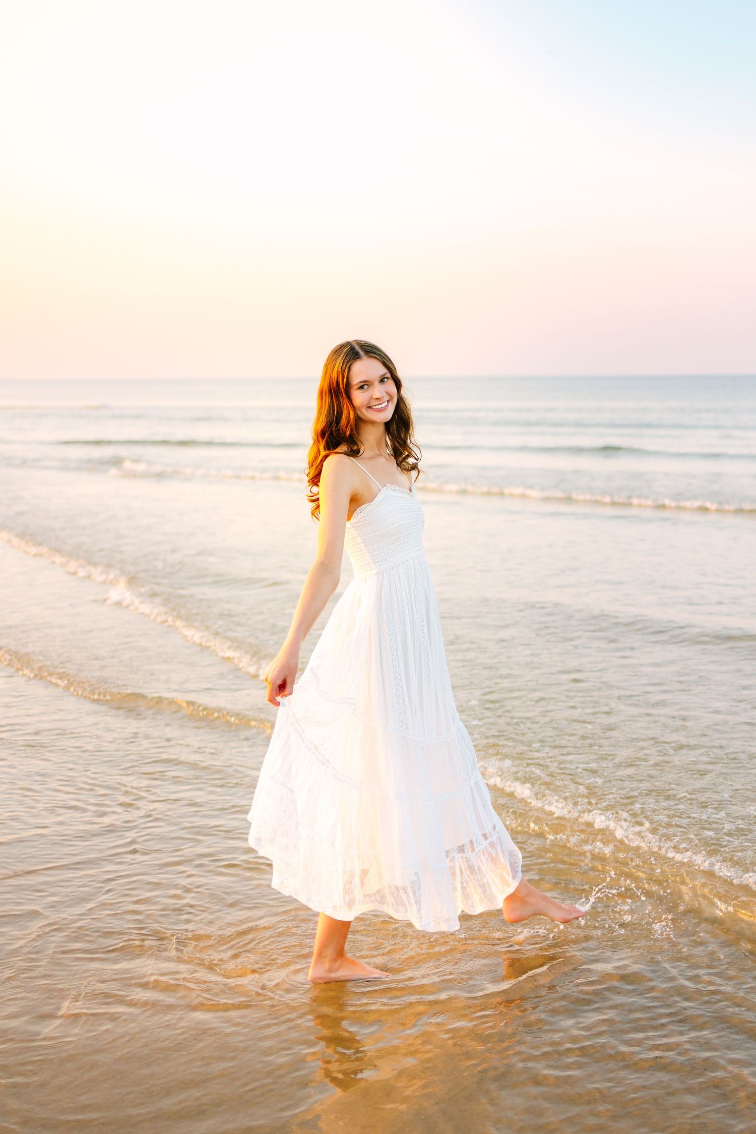 Smiling young woman in white sundress walking barefoot through shallow ocean water at golden sunset.