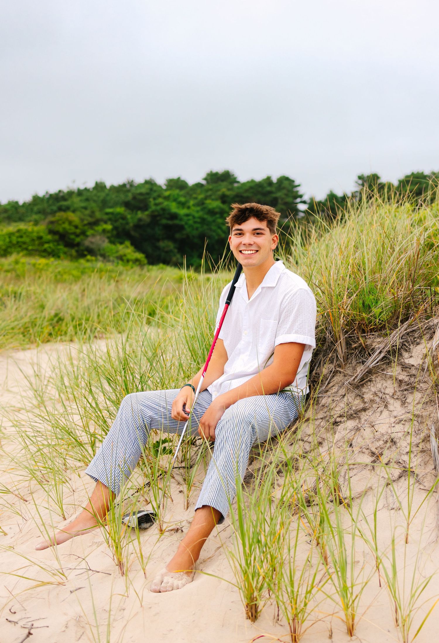 Happy young man sitting on sand dunes with golf club smiling at camera.