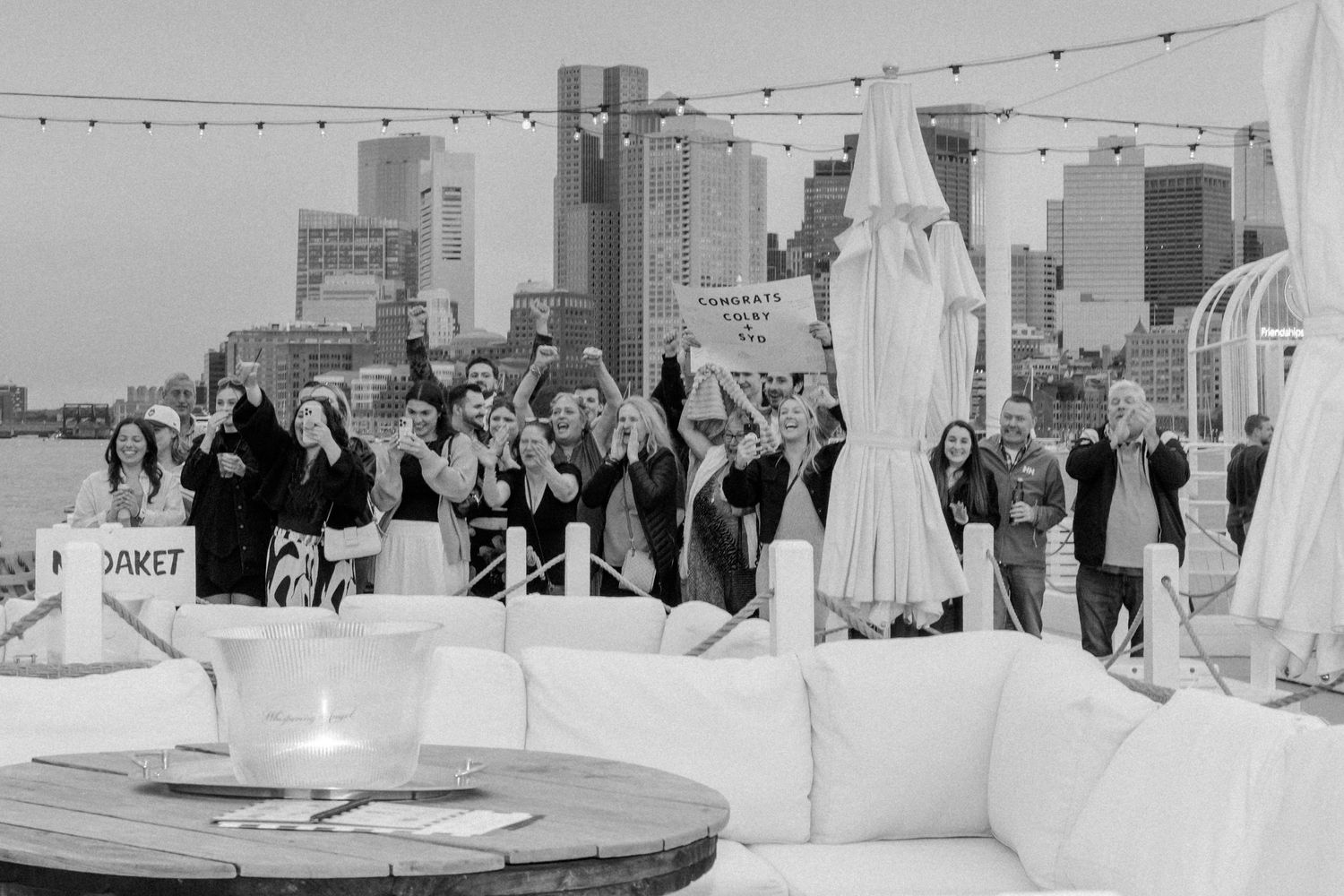 Black and white photo of a group celebrating an engagement on a boat with city skyline.