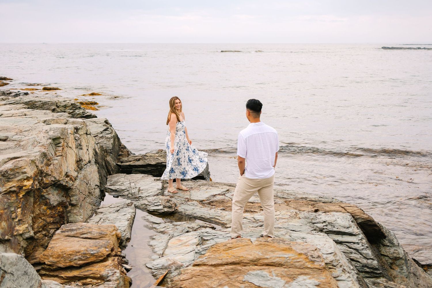Couple standing on rocky shoreline for engagement photos with man admiring his new fiancé on Ocean Drive in Newport Rhode Island