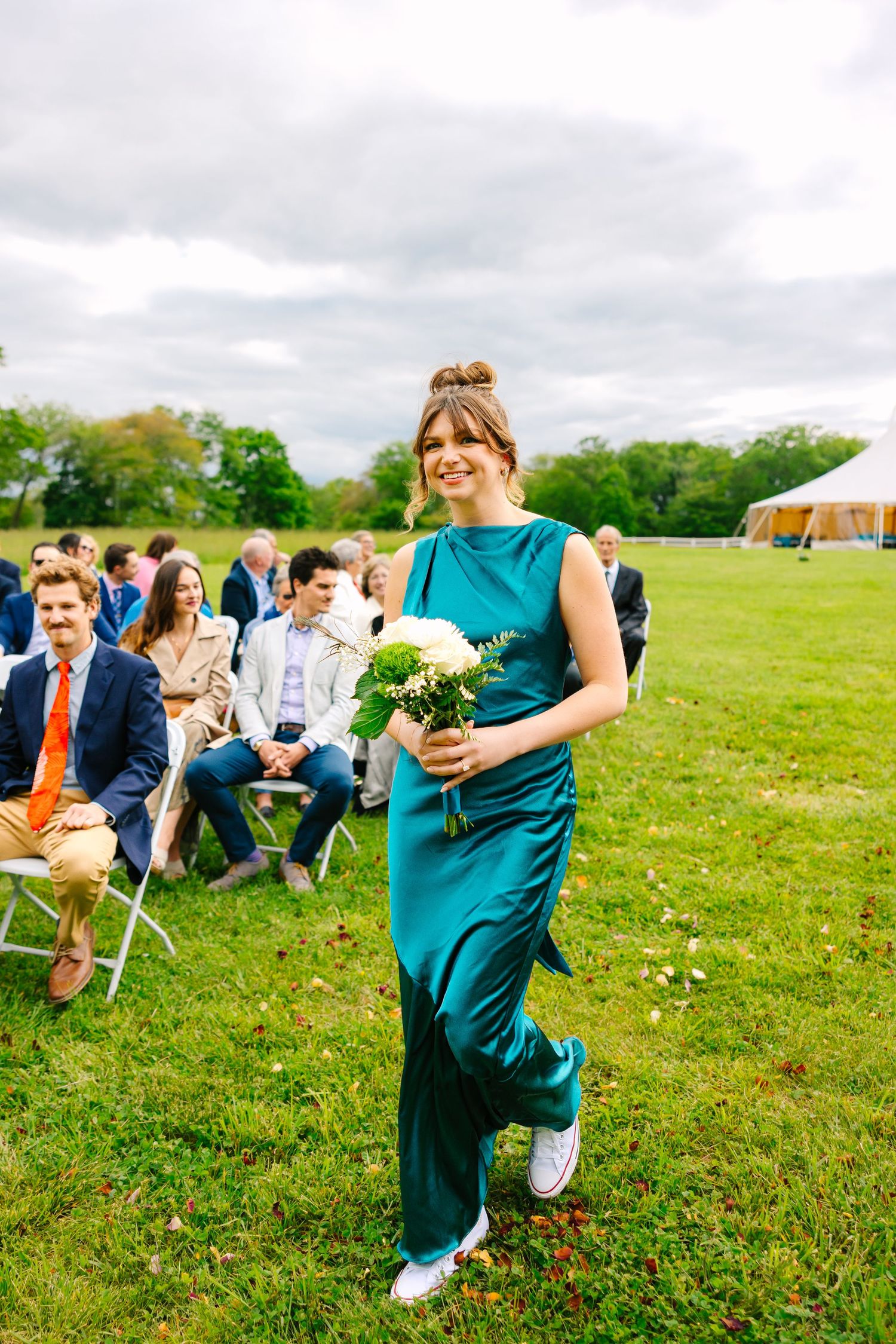 Outdoor wedding ceremony with attendees seated on white chairs in a green field as someone walks down the grassy aisle.