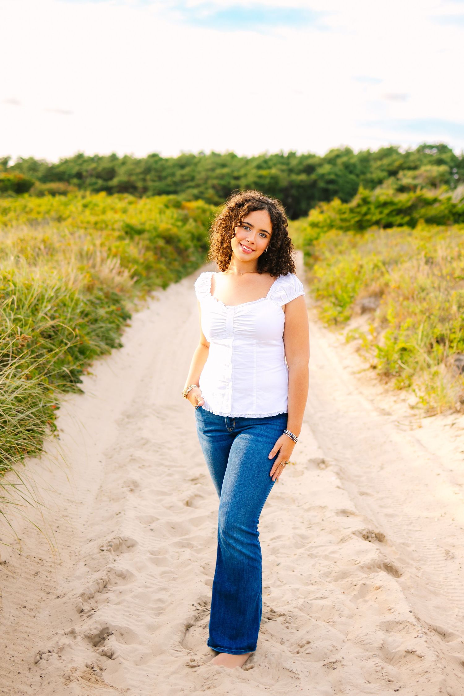 Curly-haired young woman in white top and jeans stands barefoot on sandy beach trail path.