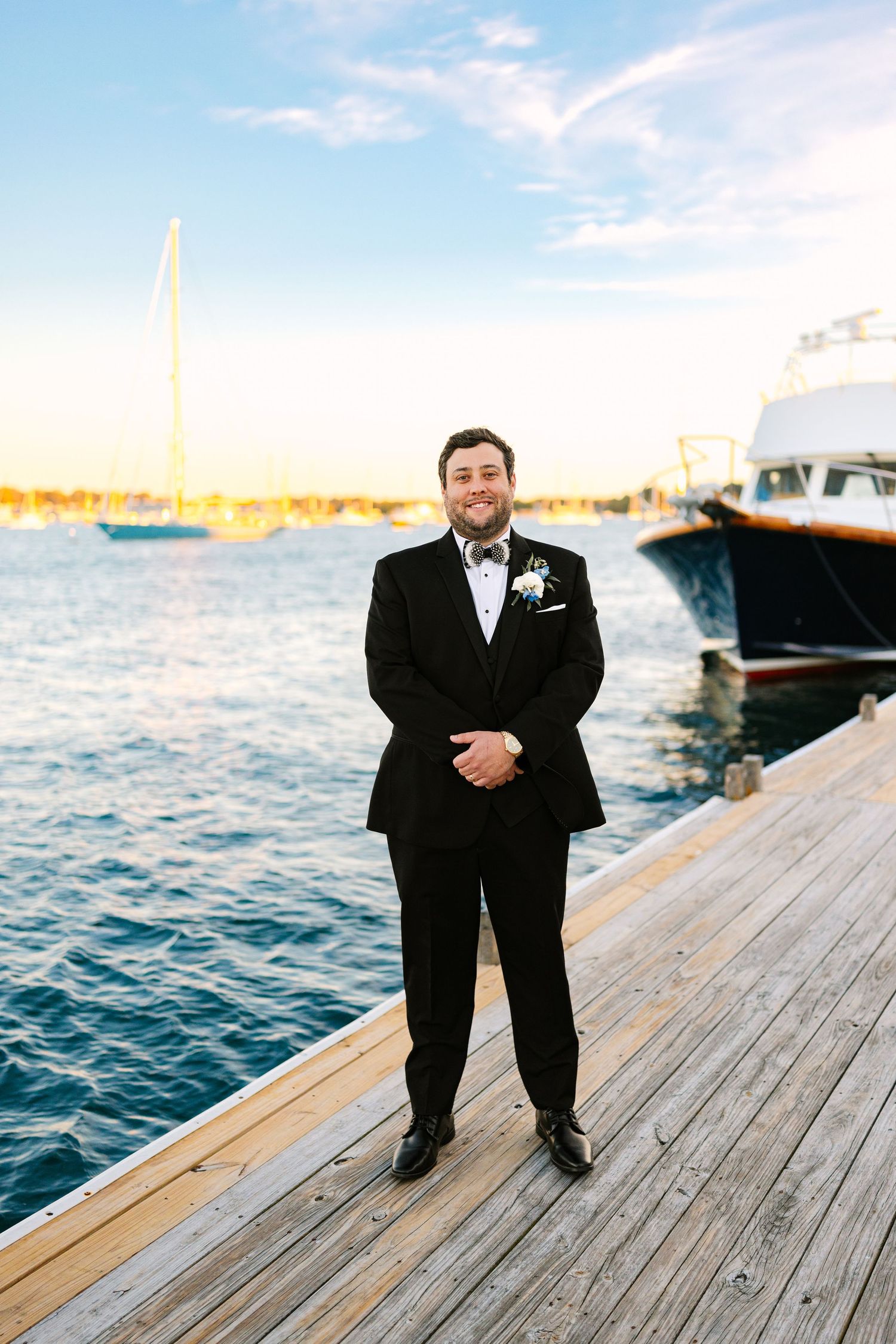 A person in a black tuxedo stands on a wooden dock by the water during sunset with boats in the background.