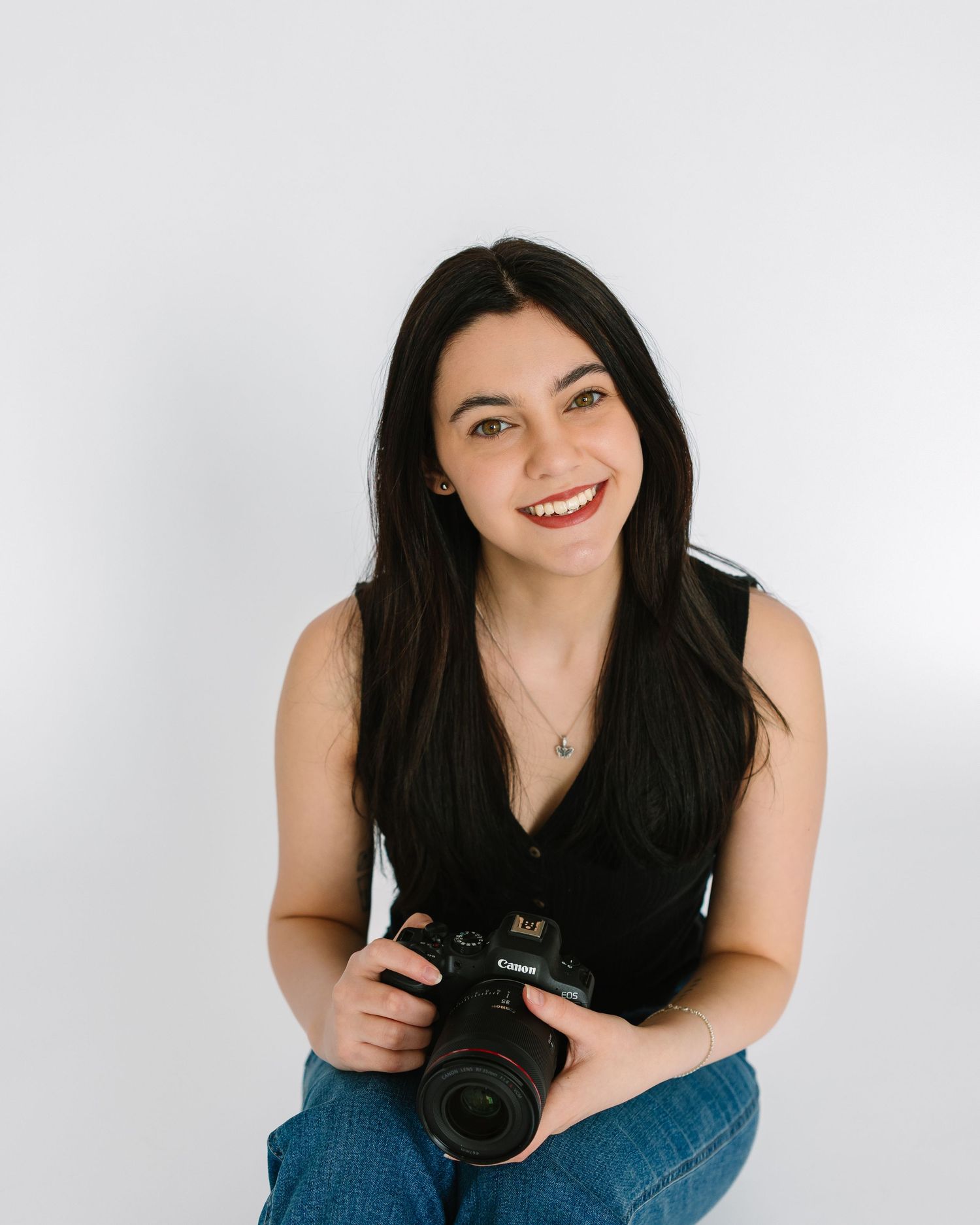 Dark-haired female photographer smiles holding a Canon camera while seated against white background.
