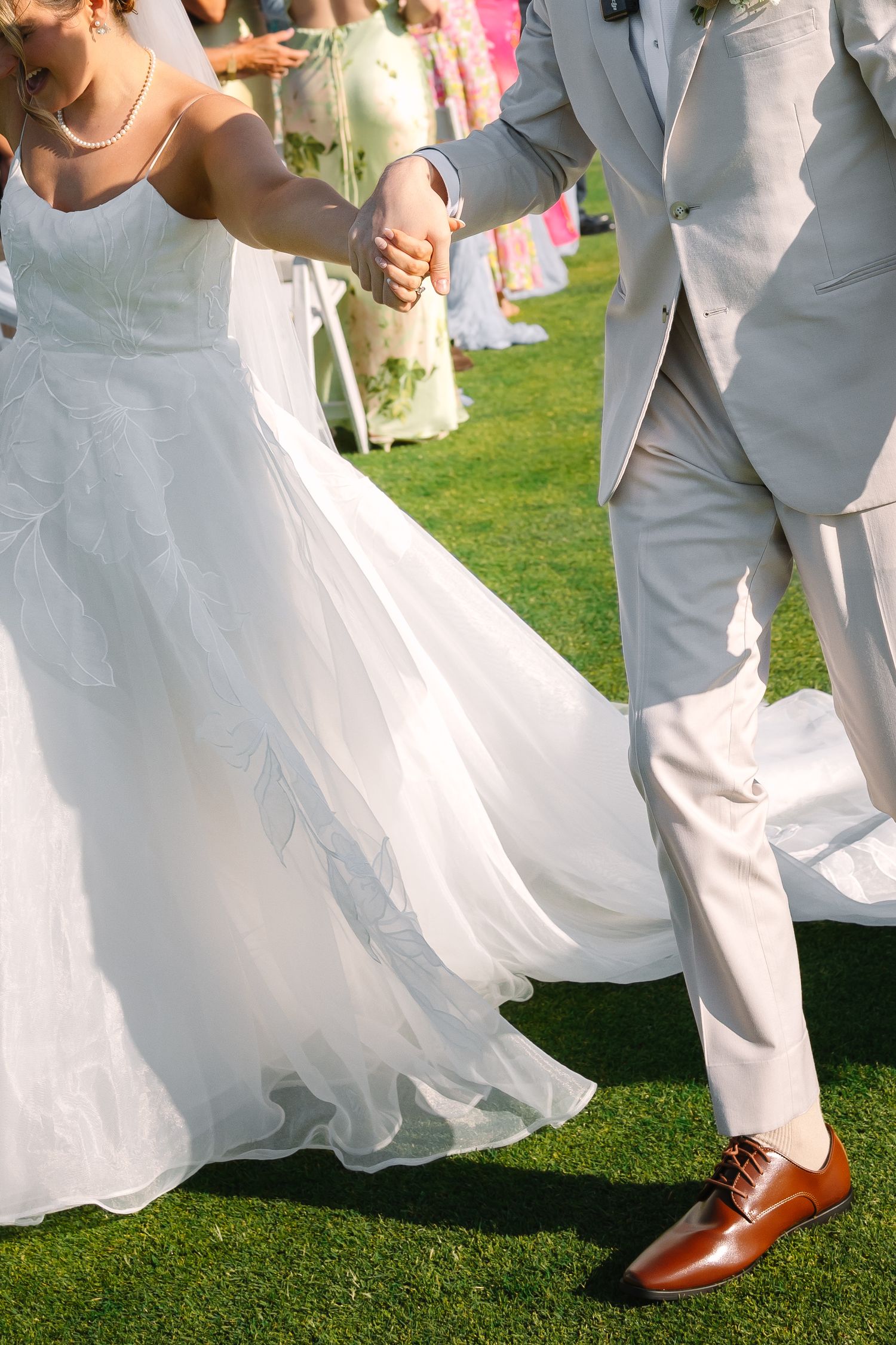 Close-up of bride in flowing white gown and groom in light gray suit holding hands while walking across lush green grass.