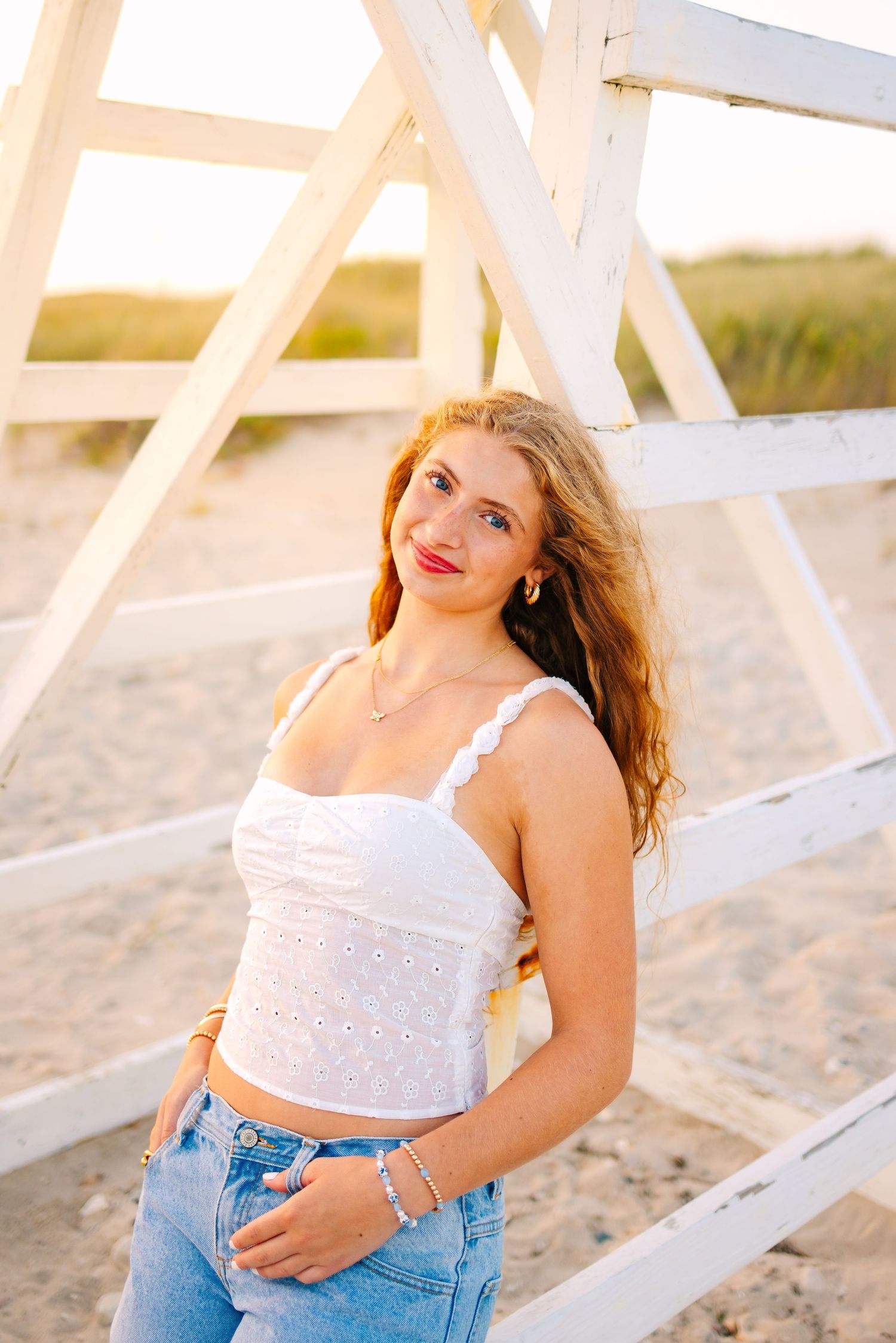 Young woman in white crop top and jeans posing by a beach lifeguard stand.