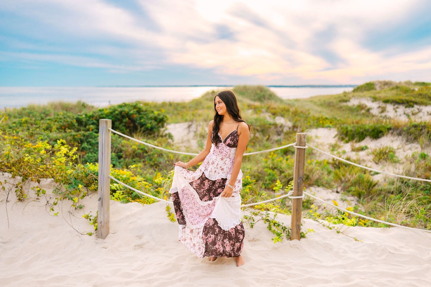 Young woman in a floral maxi dress standing on sandy beach dunes at a colorful sunset.
