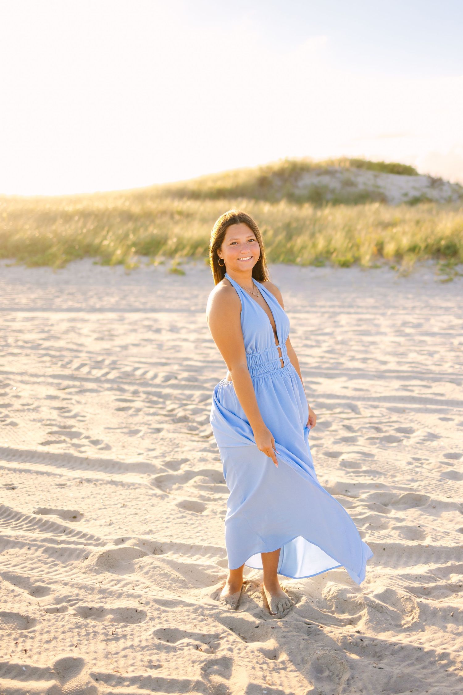 Young woman in light blue maxi dress walking on sandy beach with golden sunset dune backdrop.