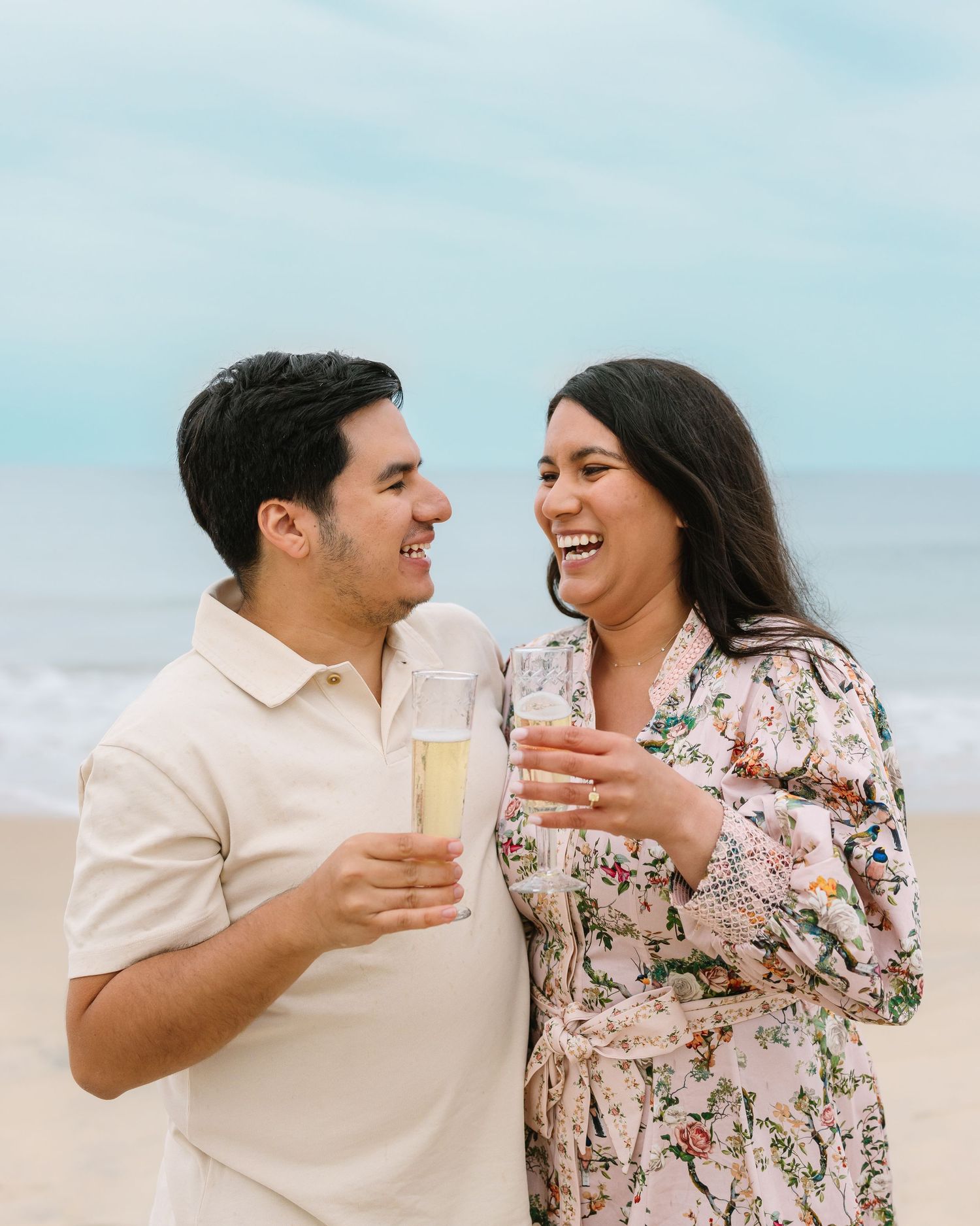 Engaged couple laughing and toasting with champagne glasses on a sandy beach.