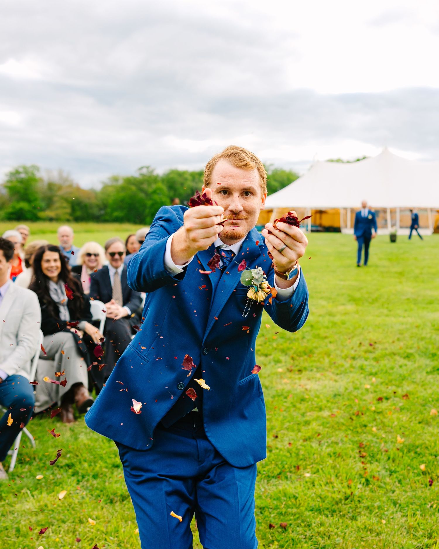 Wedding guests in blue suits raise champagne glasses for a celebratory toast on a green lawn.