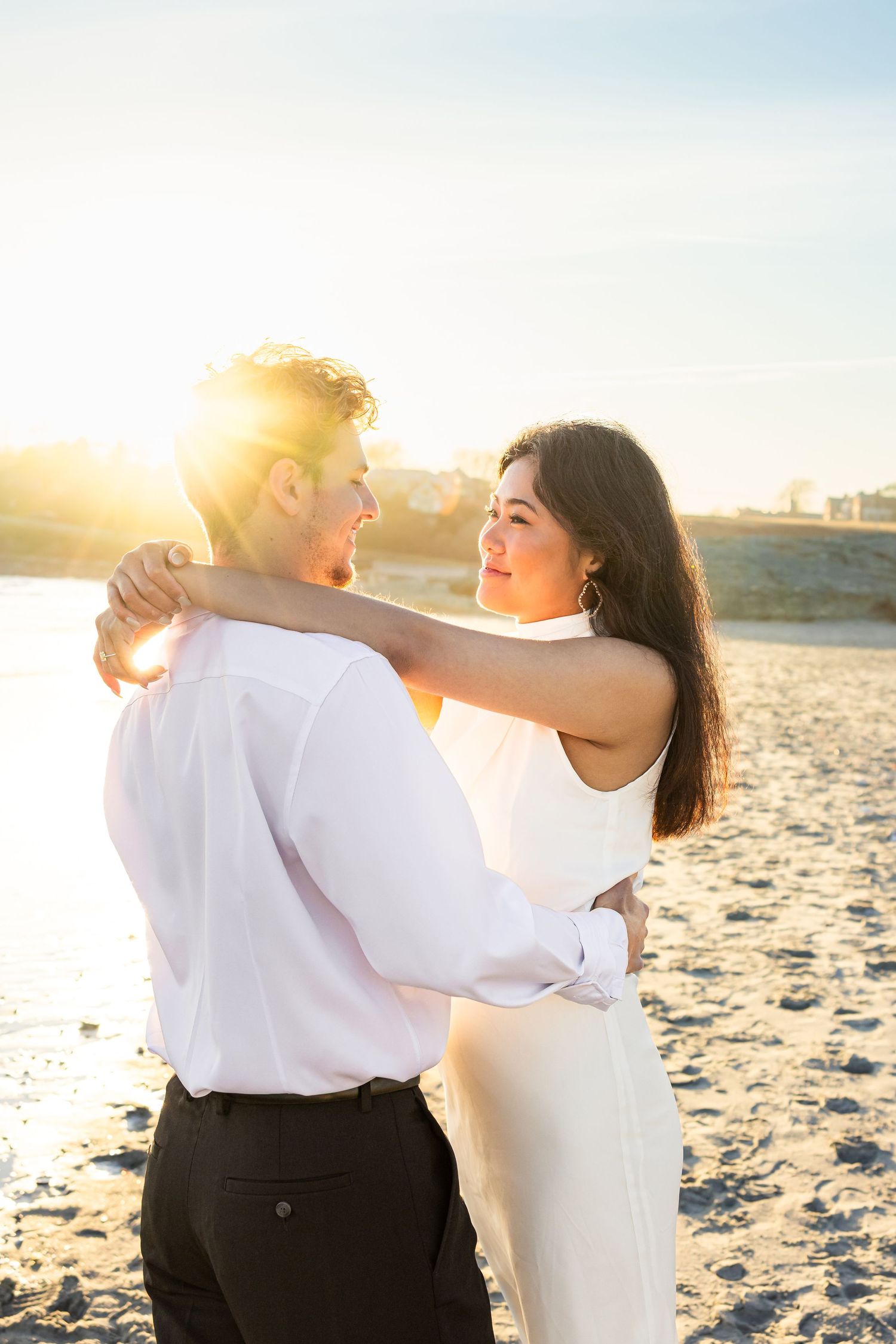 A romantic couple embraces on a sandy beach at sunset with golden sunlight glowing behind them.