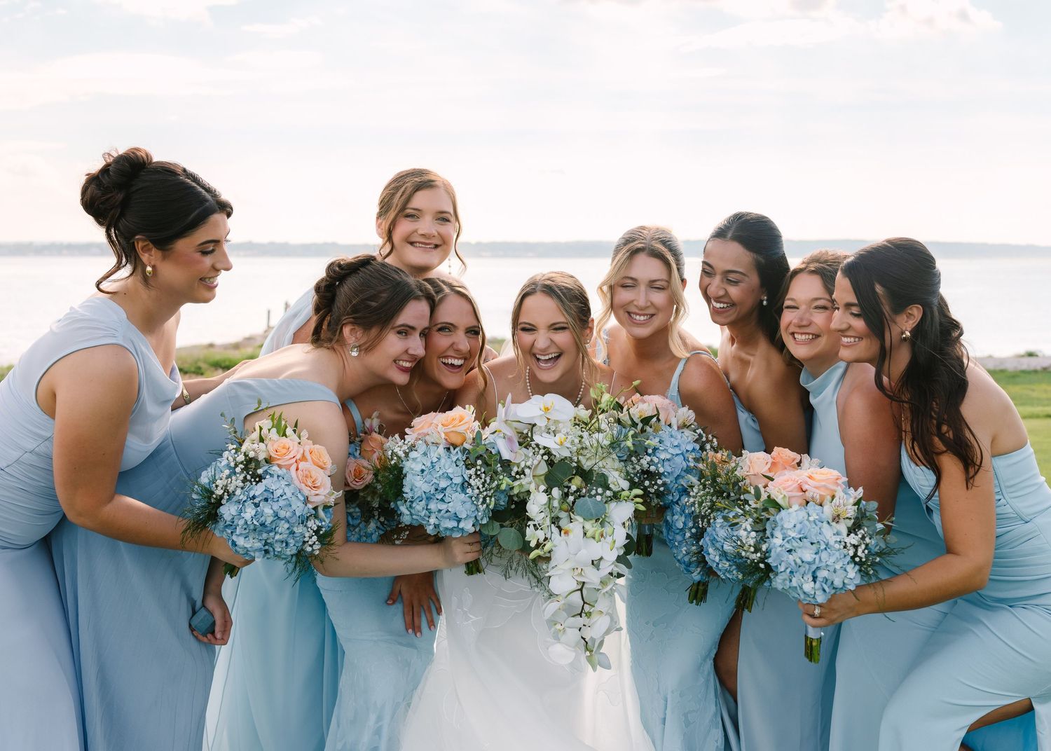 Laughing bride surrounded by bridesmaids in blue dresses holding hydrangea bouquets near waterfront.