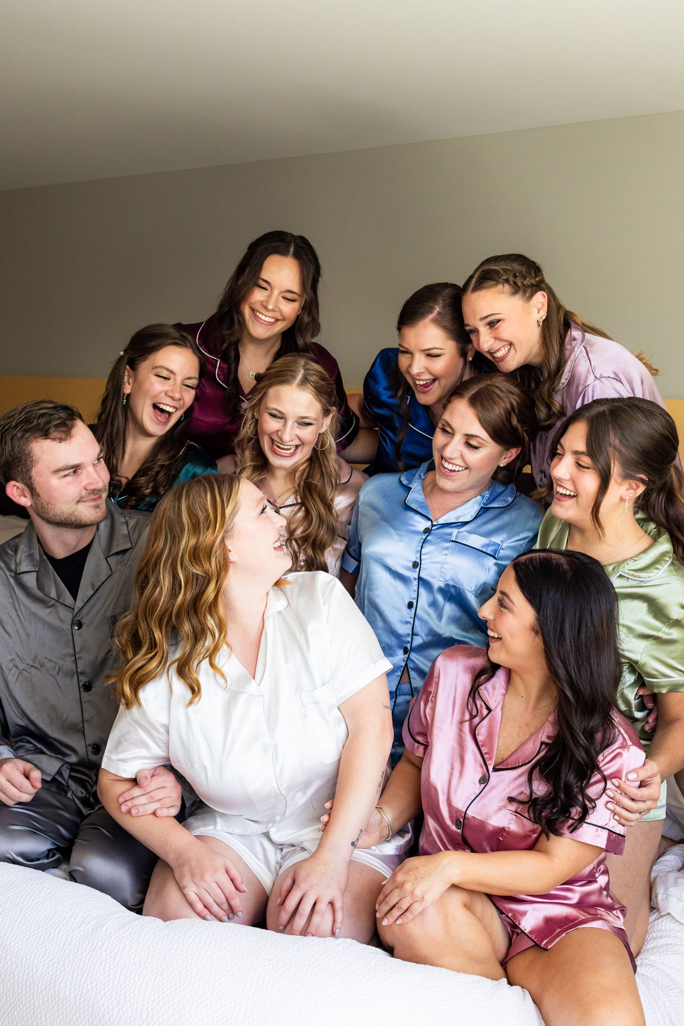 Bride in white satin robe laughing joyfully surrounded by bridesmaids in colorful satin pajamas on bed.