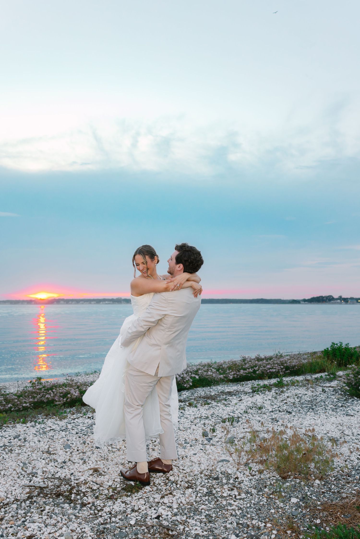 Groom lifts smiling bride at the rocky shoreline during a breathtaking pink and blue sunset over the calm ocean waters.