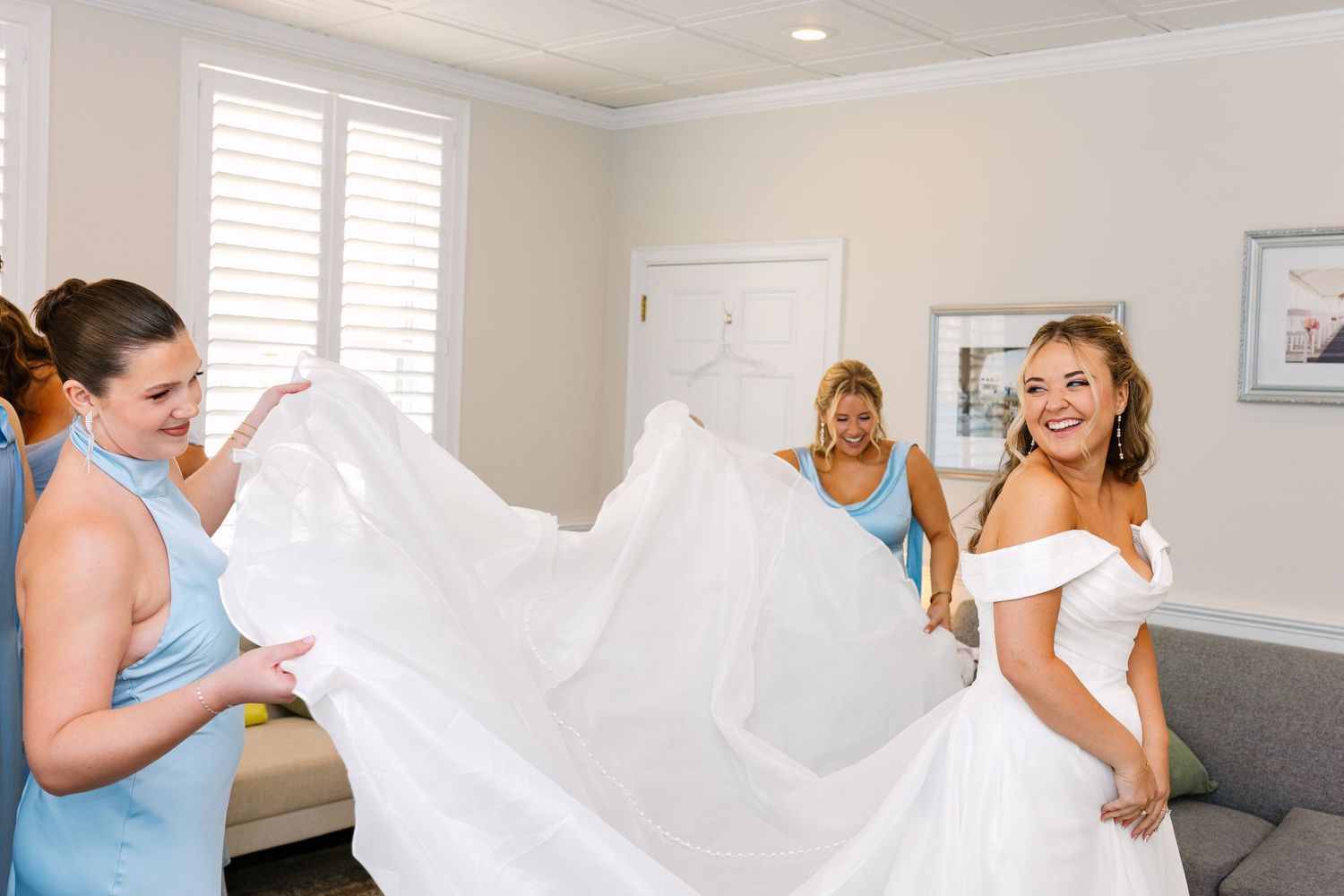 Wedding party helps bride get ready in bright white room with bridesmaids assisting with dress and preparations at The Regatta Place in RI