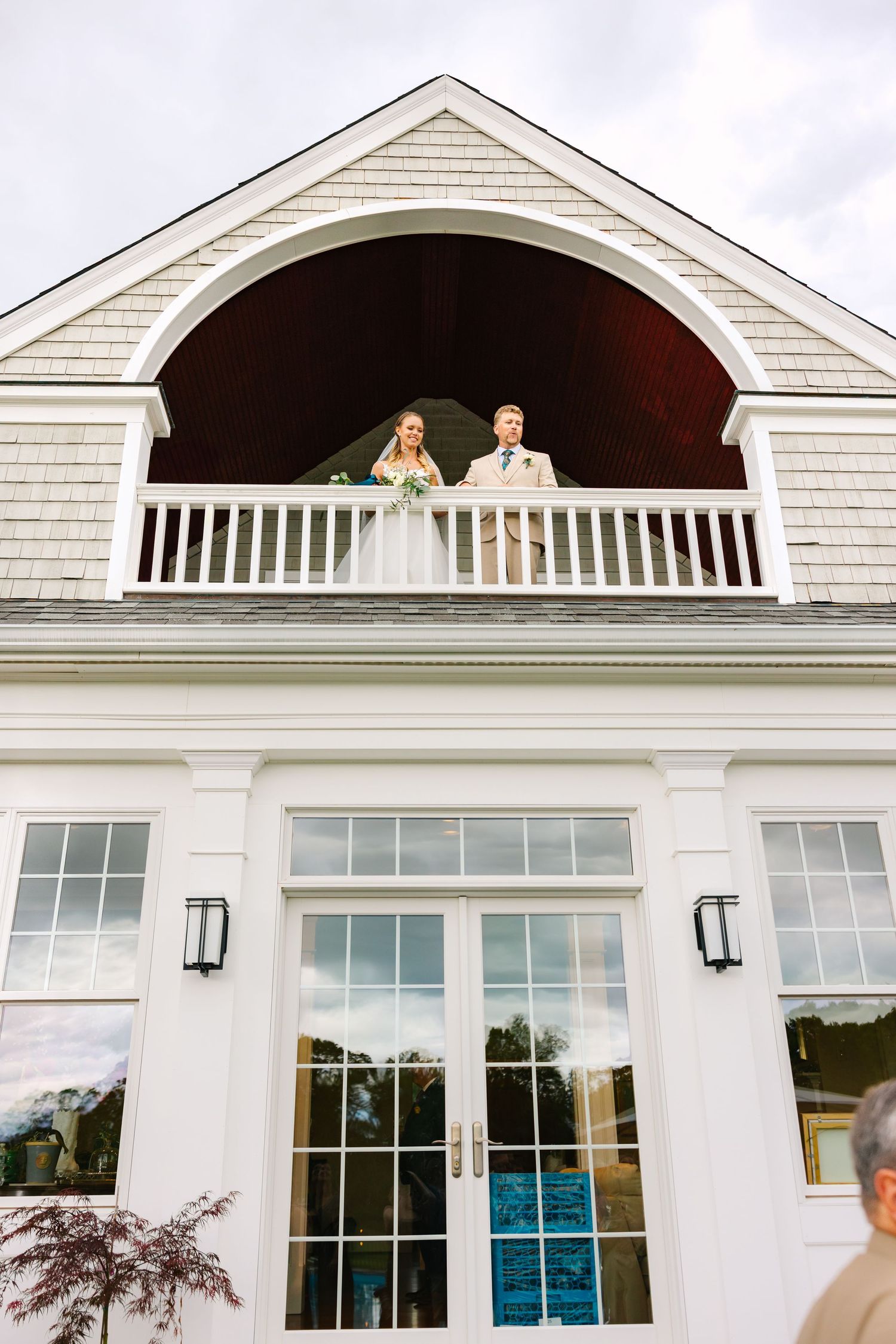A white wedding venue with large windows and architectural details shows a couple posing in the upper arched window.