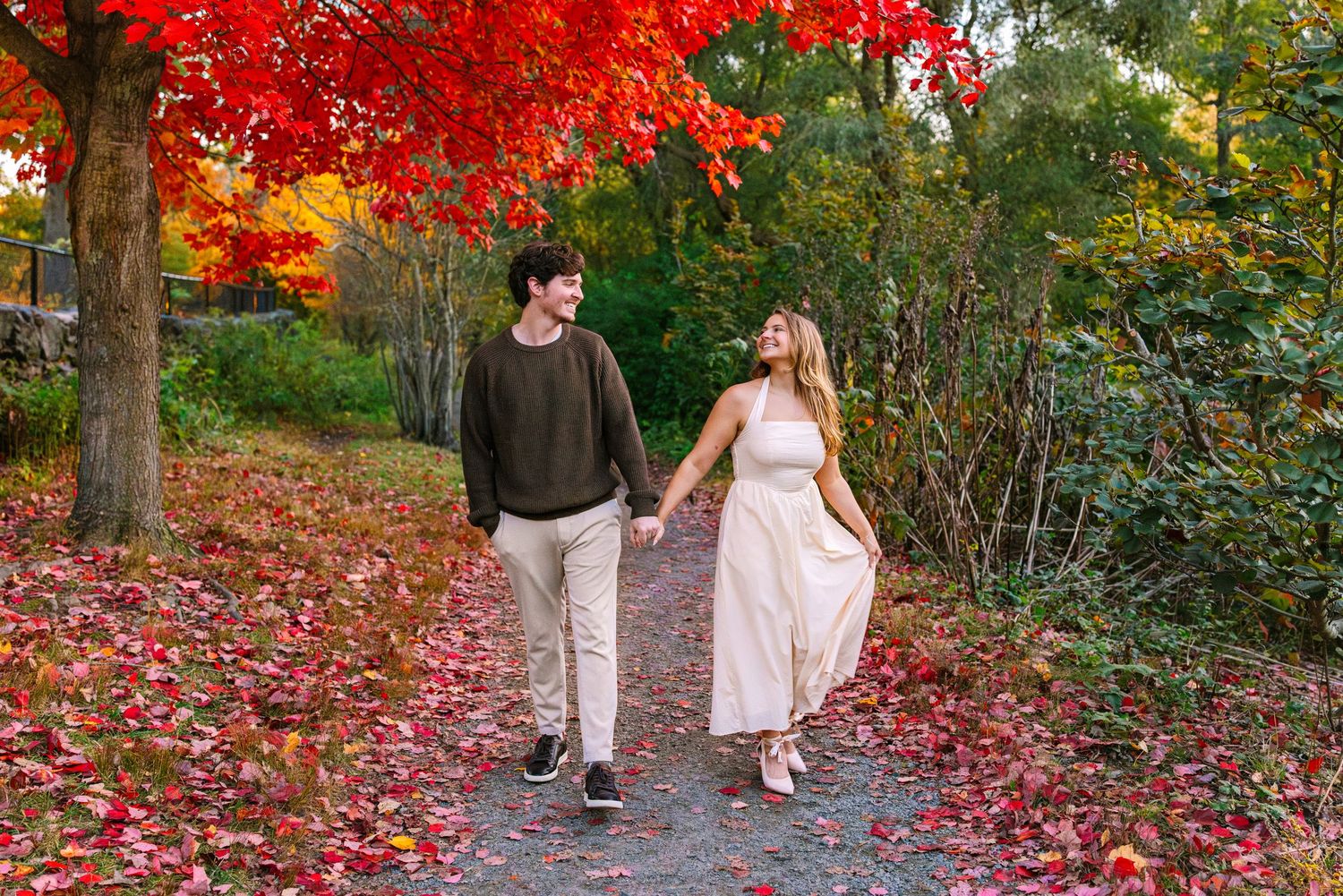 Two people walk along a scenic autumn path covered in vibrant red fallen leaves during fall season.