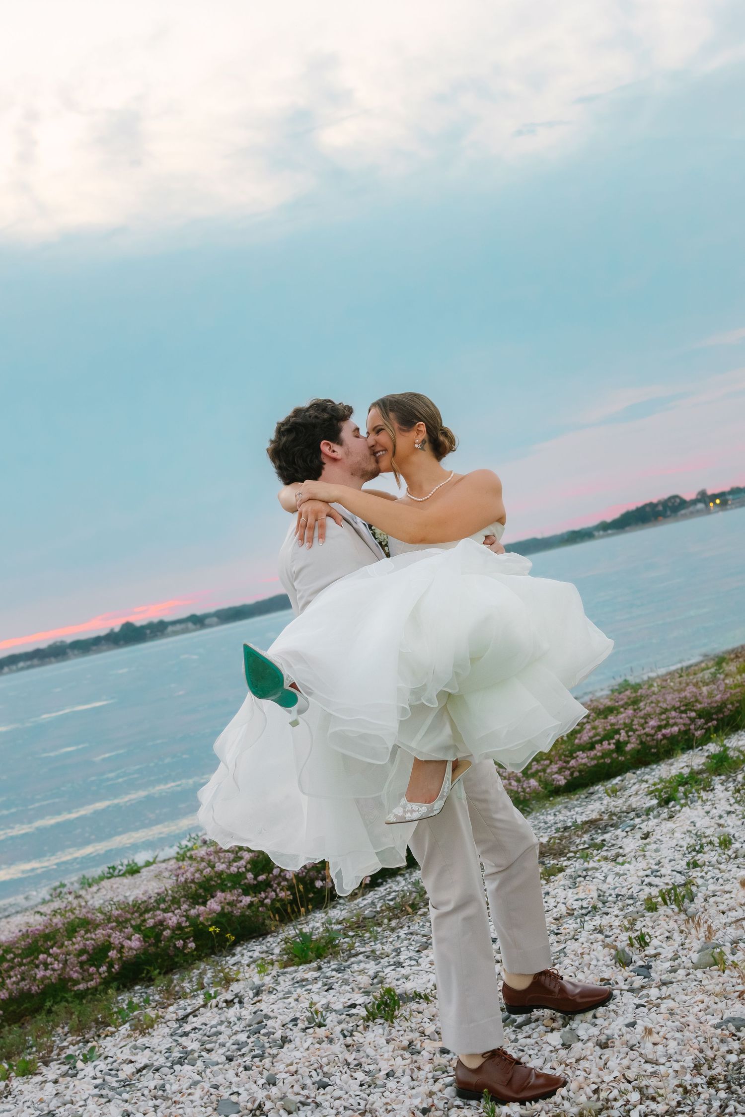 Groom lifting bride in white dress for a kiss on rocky shoreline at dusk at Warwick Country Club in Rhode Island.