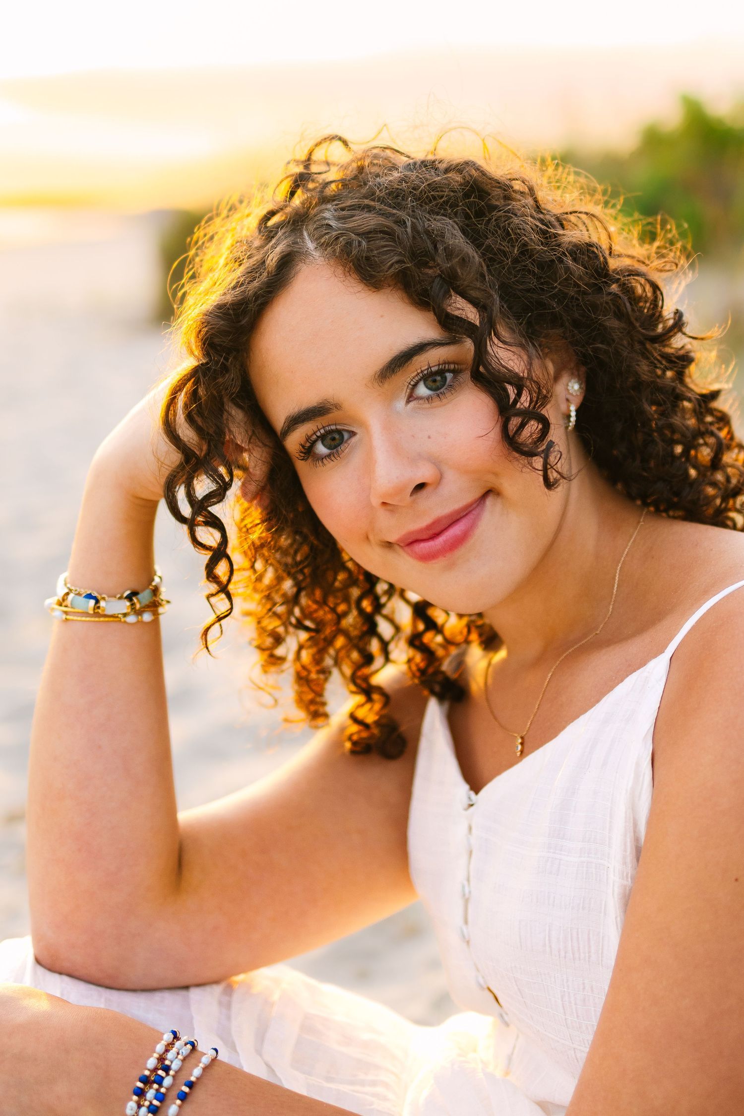 Close-up portrait of curly-haired young woman with bracelets smiling at golden hour beach.