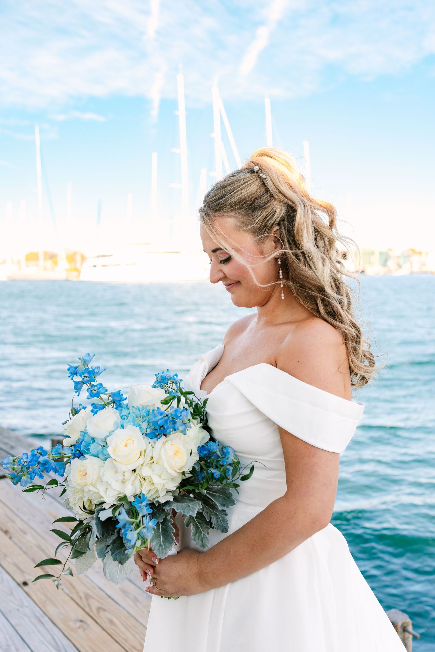 Bride in off-shoulder white dress holds stunning blue and white bouquet while standing on dock by ocean.