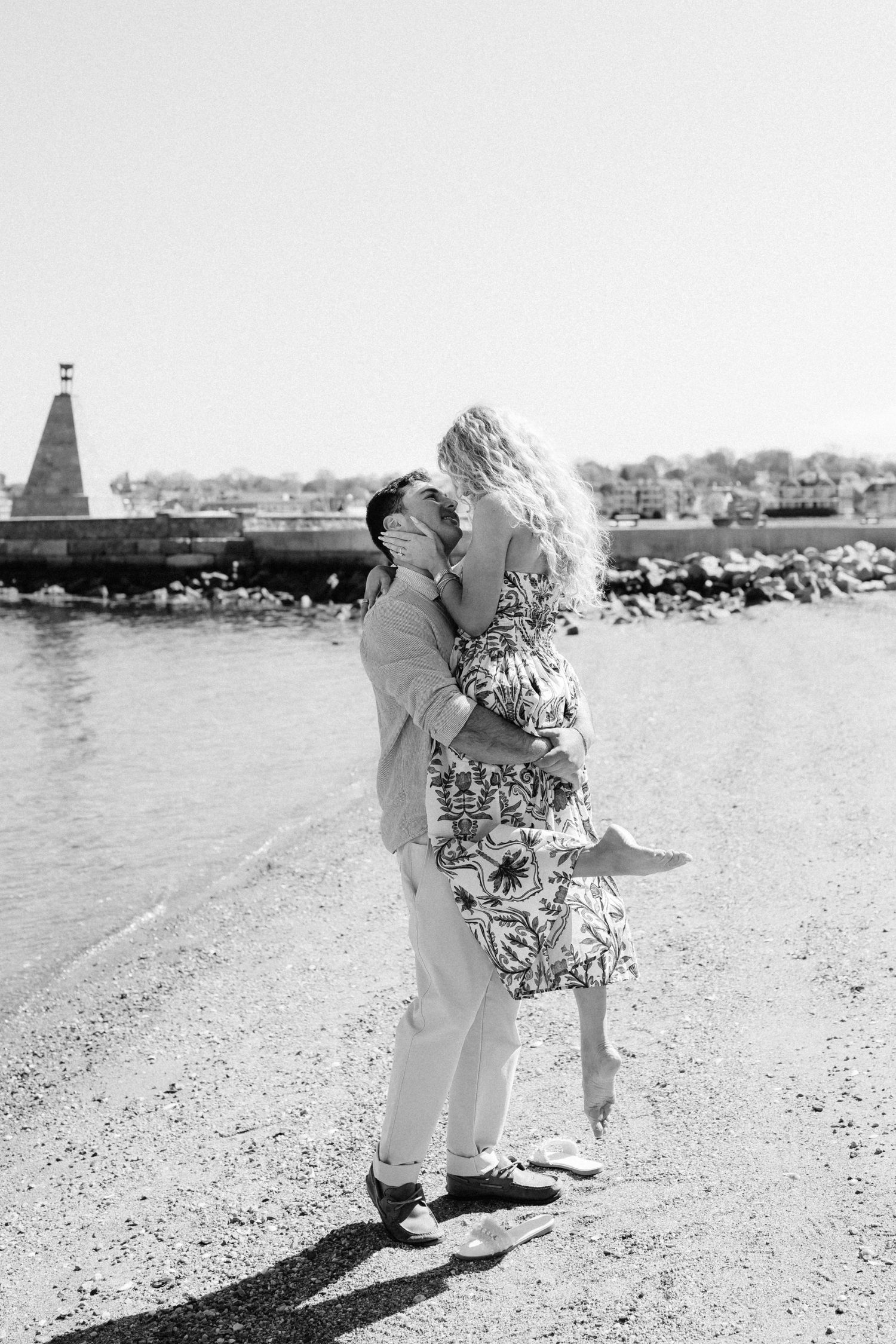 Black and white photo of couple embracing and kissing on a rocky beach shoreline near the water.