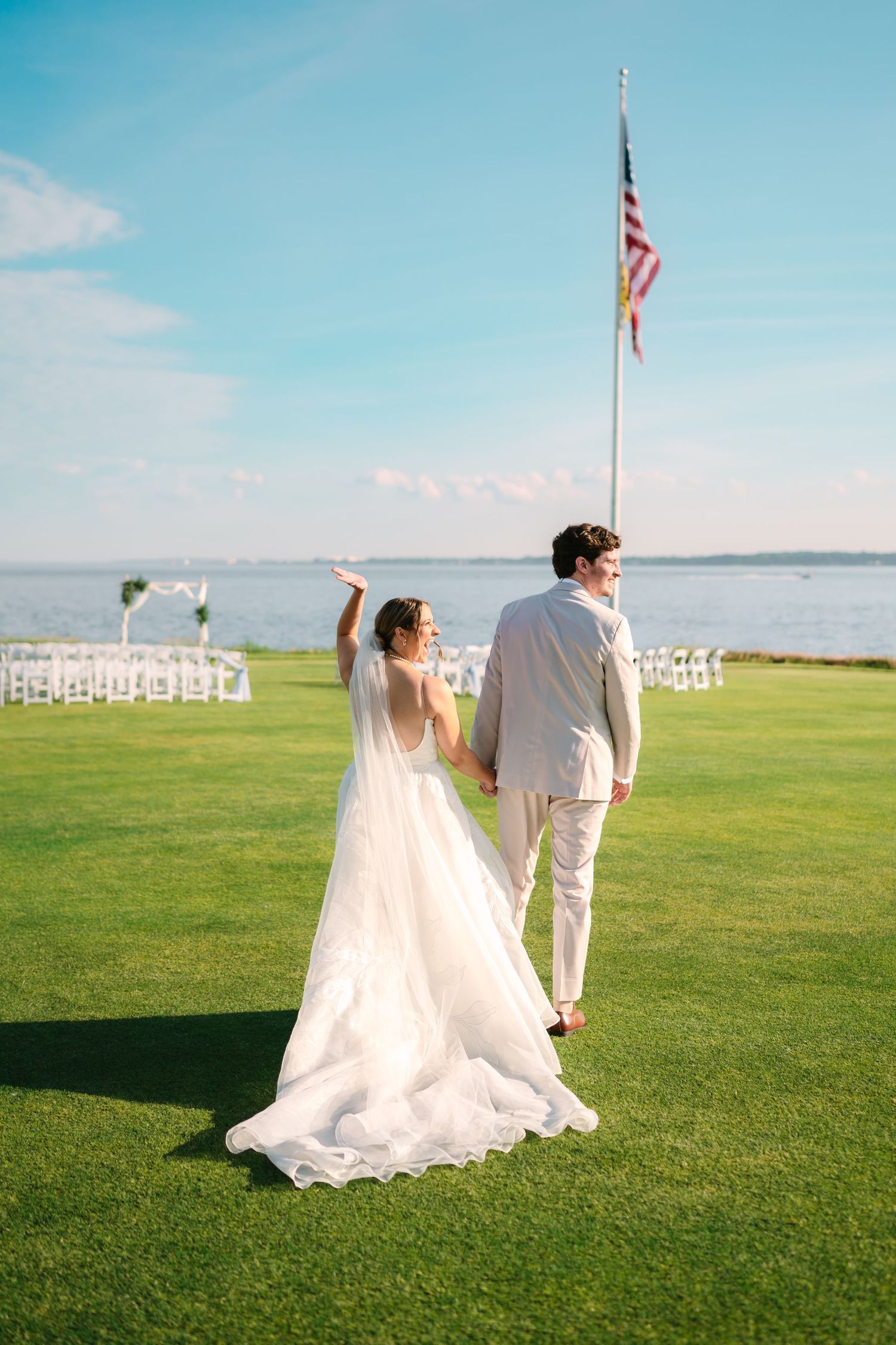 A bride and groom walk hand in hand across a lush green lawn toward a waterfront wedding venue.