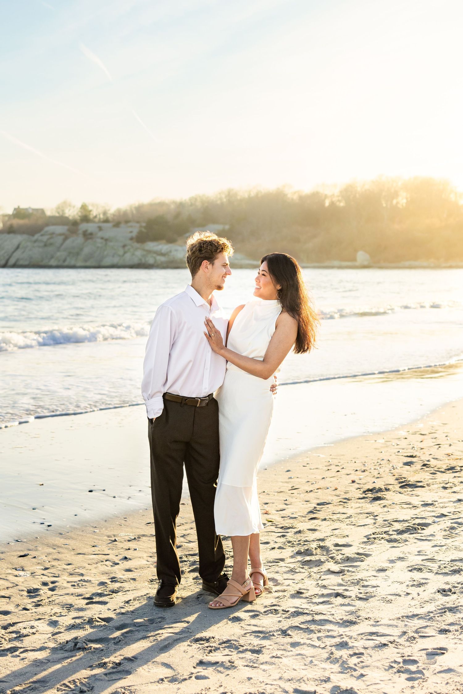 A couple shares a romantic moment at sunset on a sandy beach with cliffs in the background.