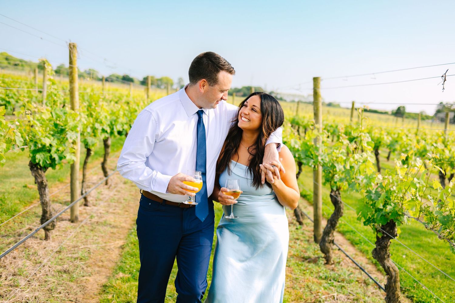 A couple embrace in a scenic vineyard with grapevines and rolling hills during a sunny afternoon.